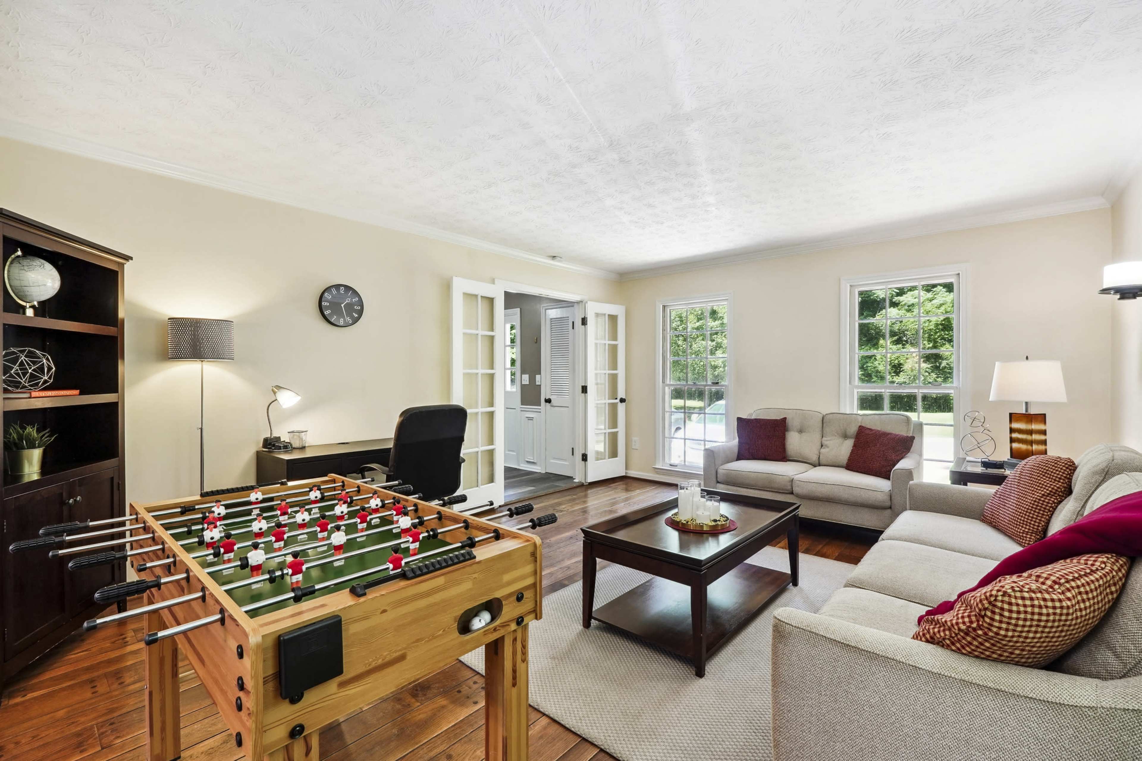 The image shows a living room featuring a foosball table, a beige sofa, a dark wood coffee table, and large windows that let in natural light.
