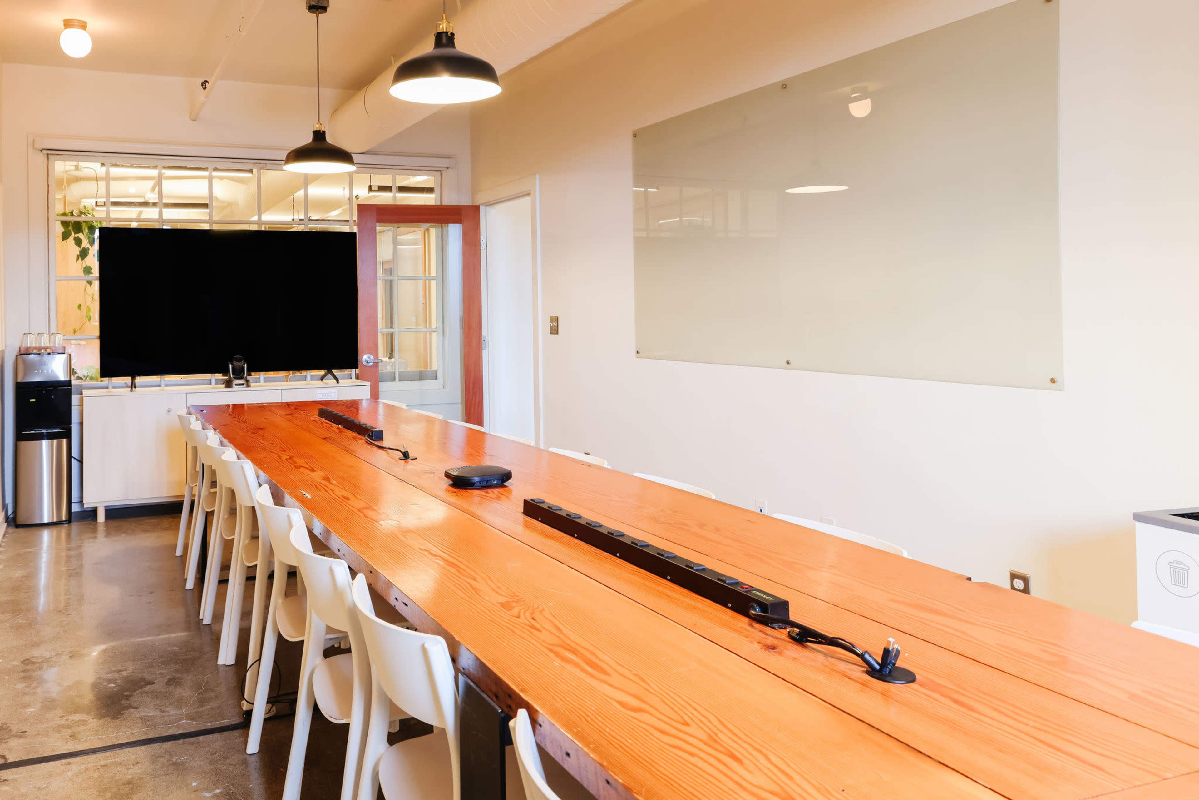 A long wooden conference table with white chairs is positioned in a well-lit meeting room featuring a large screen at one end and a clear whiteboard on the wall.