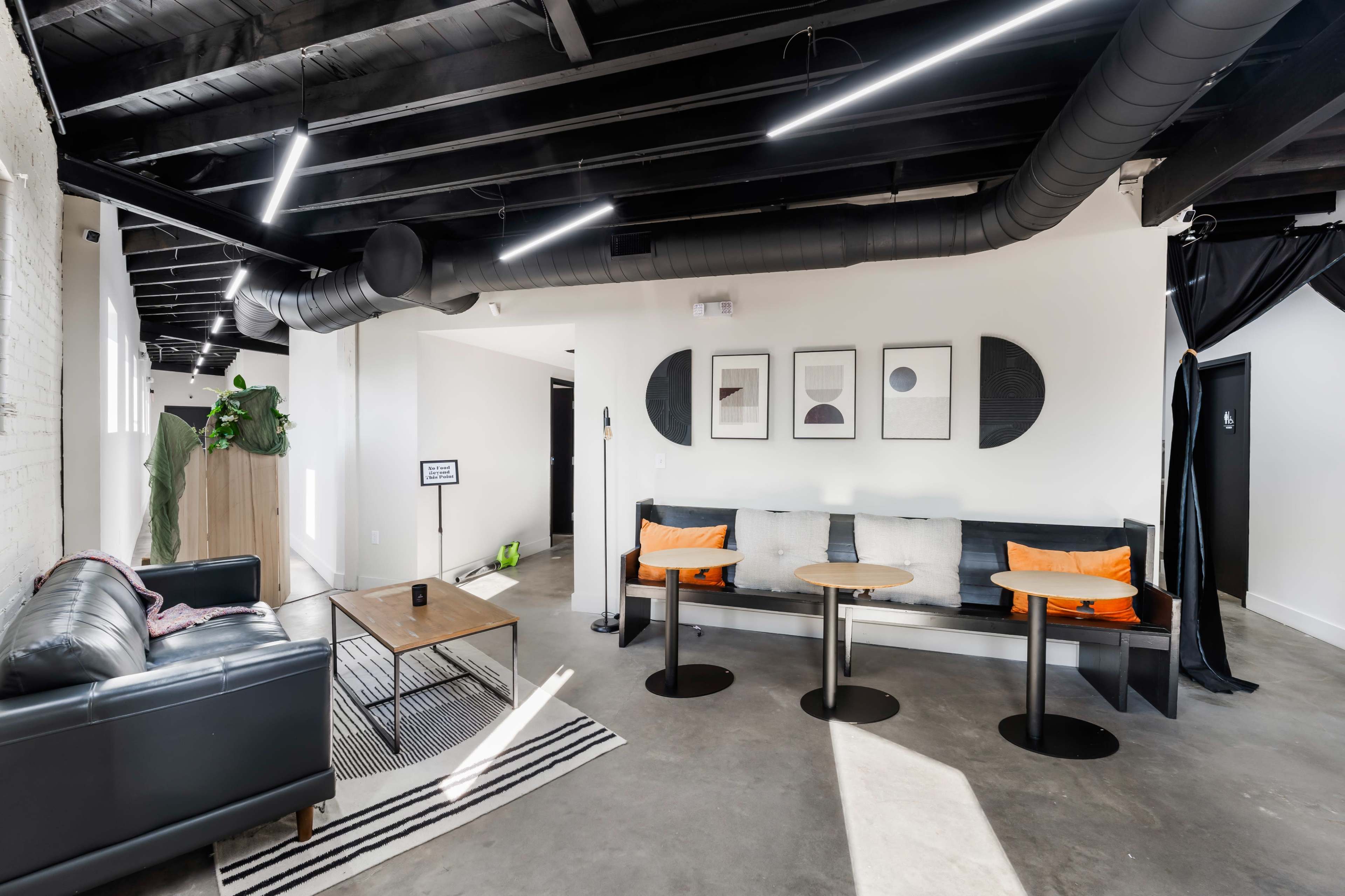 The image shows a modern interior space featuring a seating area with a black leather couch, two wooden tables, and decorative framed artwork on the walls.