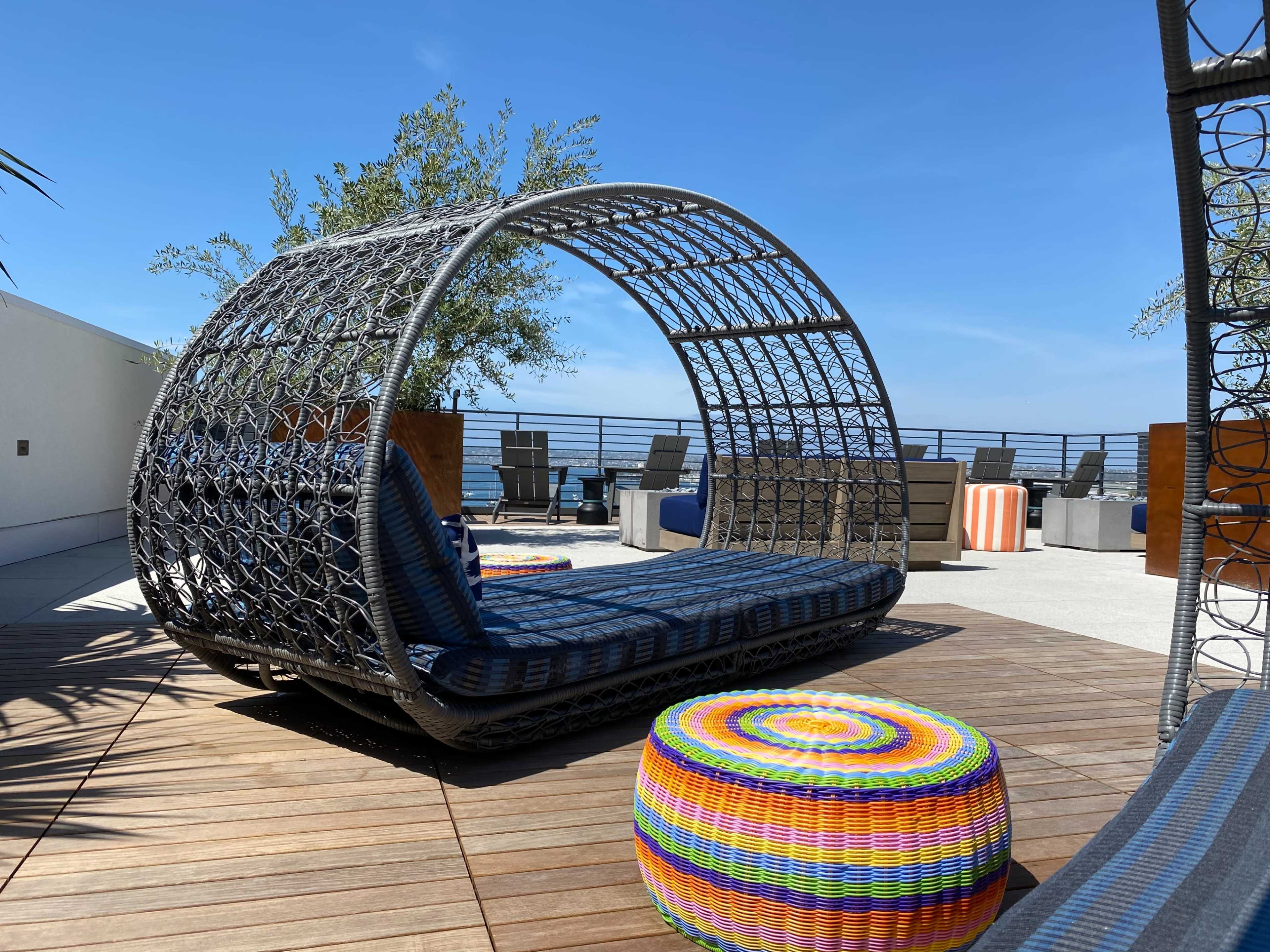 The image shows a modern outdoor seating area featuring a large circular swing chair and a colorful woven table on a sunlit patio overlooking the ocean.