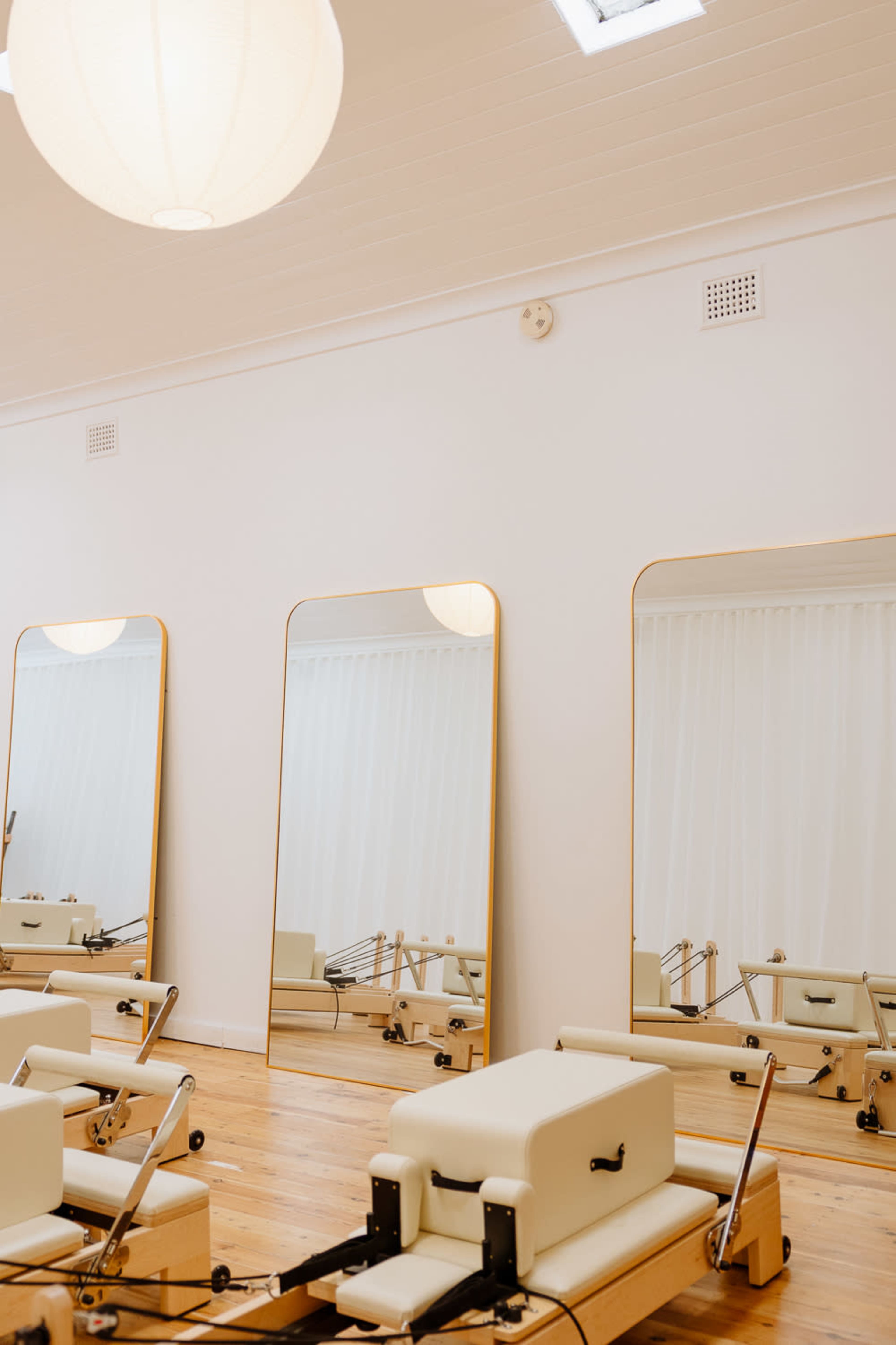The image shows a bright room with wooden flooring, featuring three large, framed mirrors and a pilates reformer machine in the foreground.