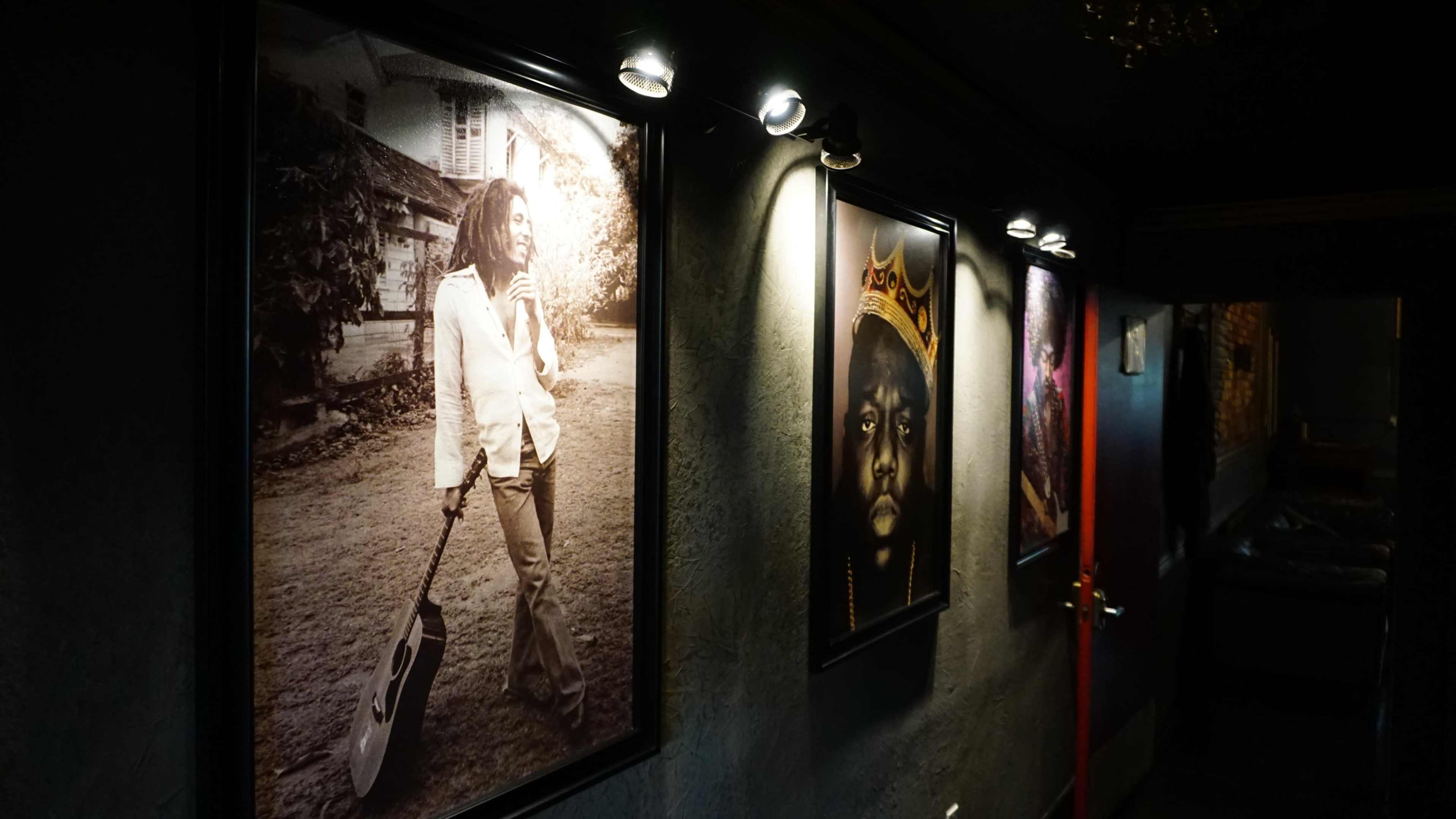 The image shows a dimly lit hallway adorned with framed black-and-white photographs of musicians, featuring one holding a guitar and another wearing a crown.
