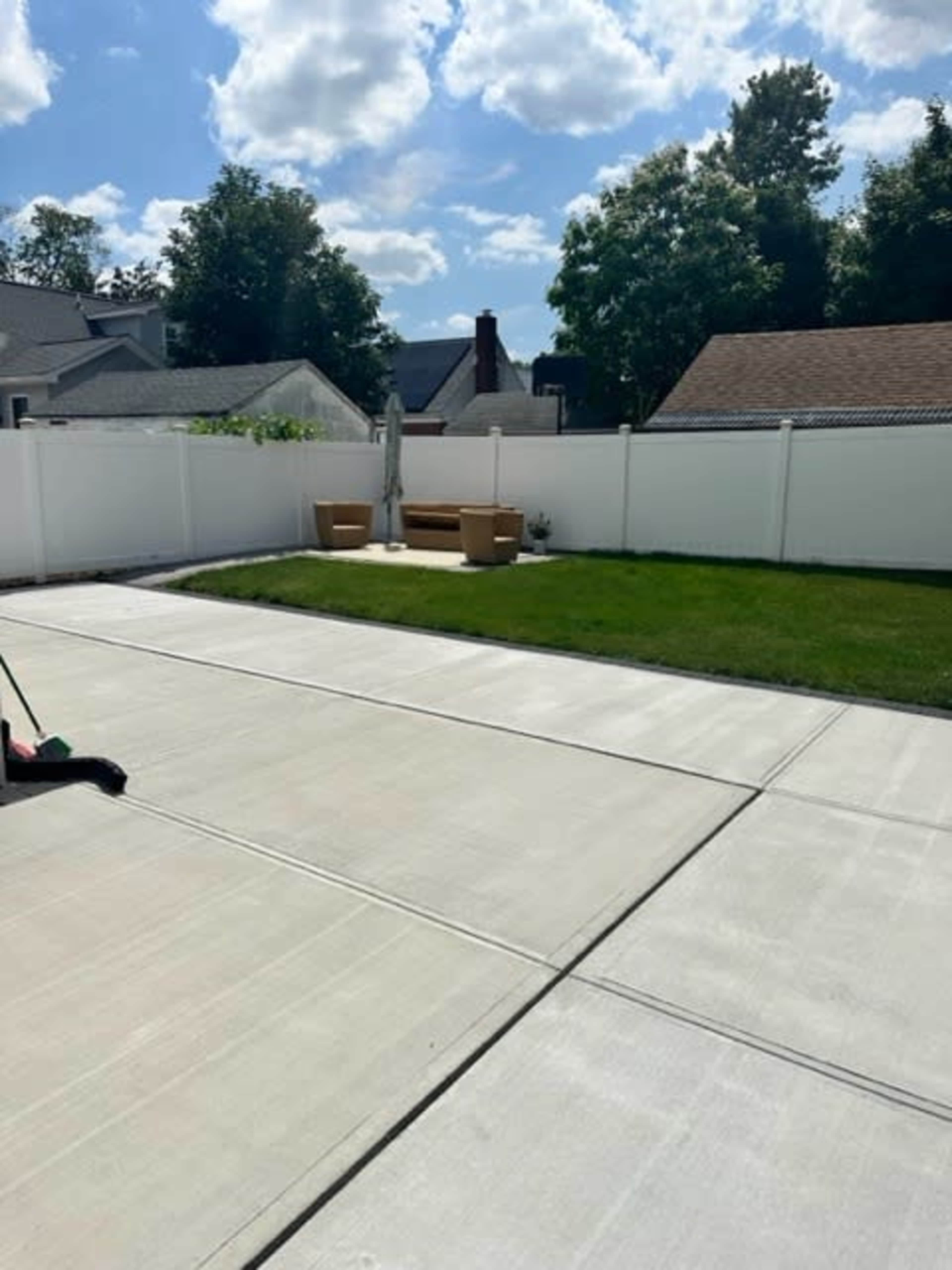 A concrete patio with a green lawn and a seating area is enclosed by a white fence under a partly cloudy sky.