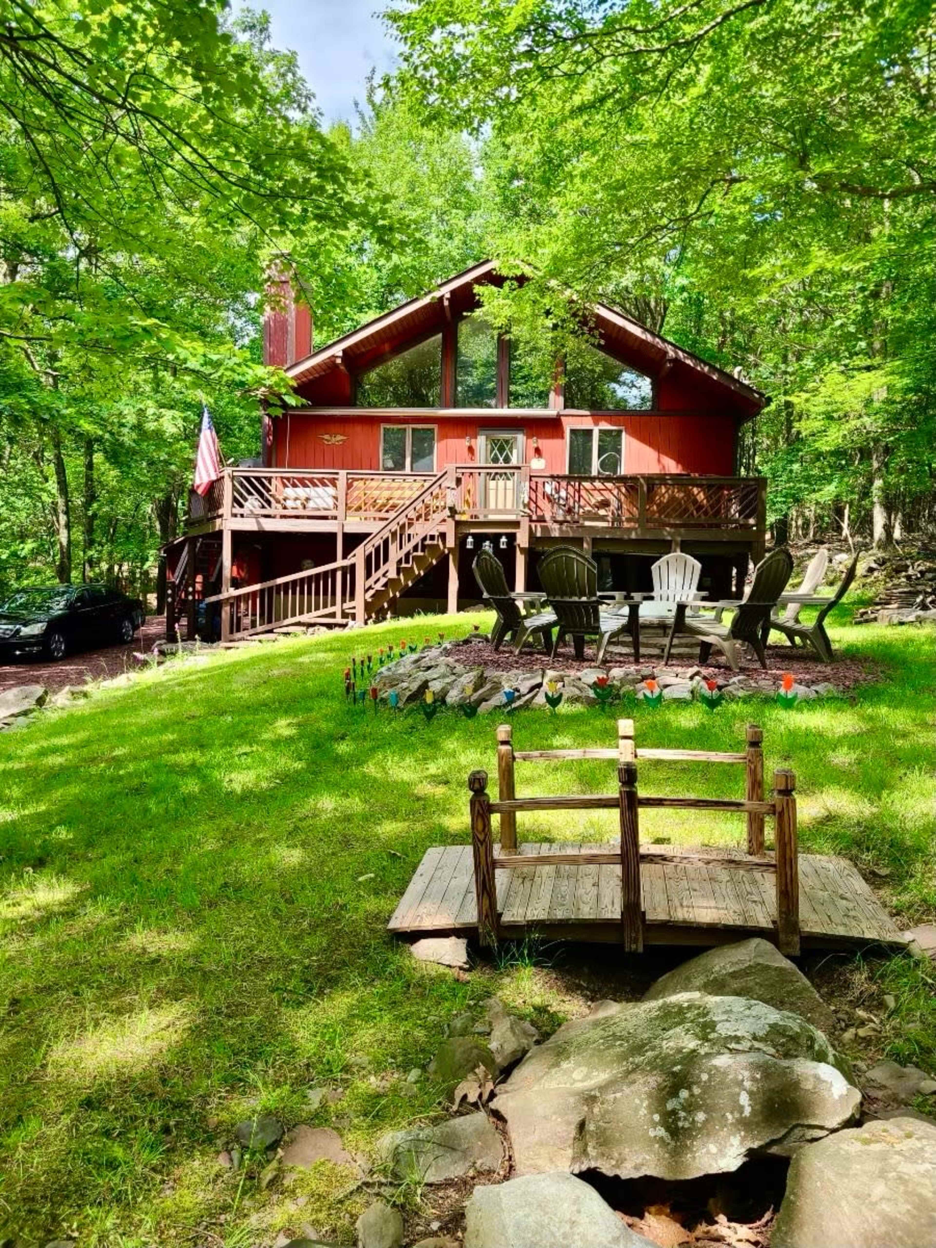 A red cabin is surrounded by greenery, featuring a deck with chairs and a small wooden bridge leading to a stone area.