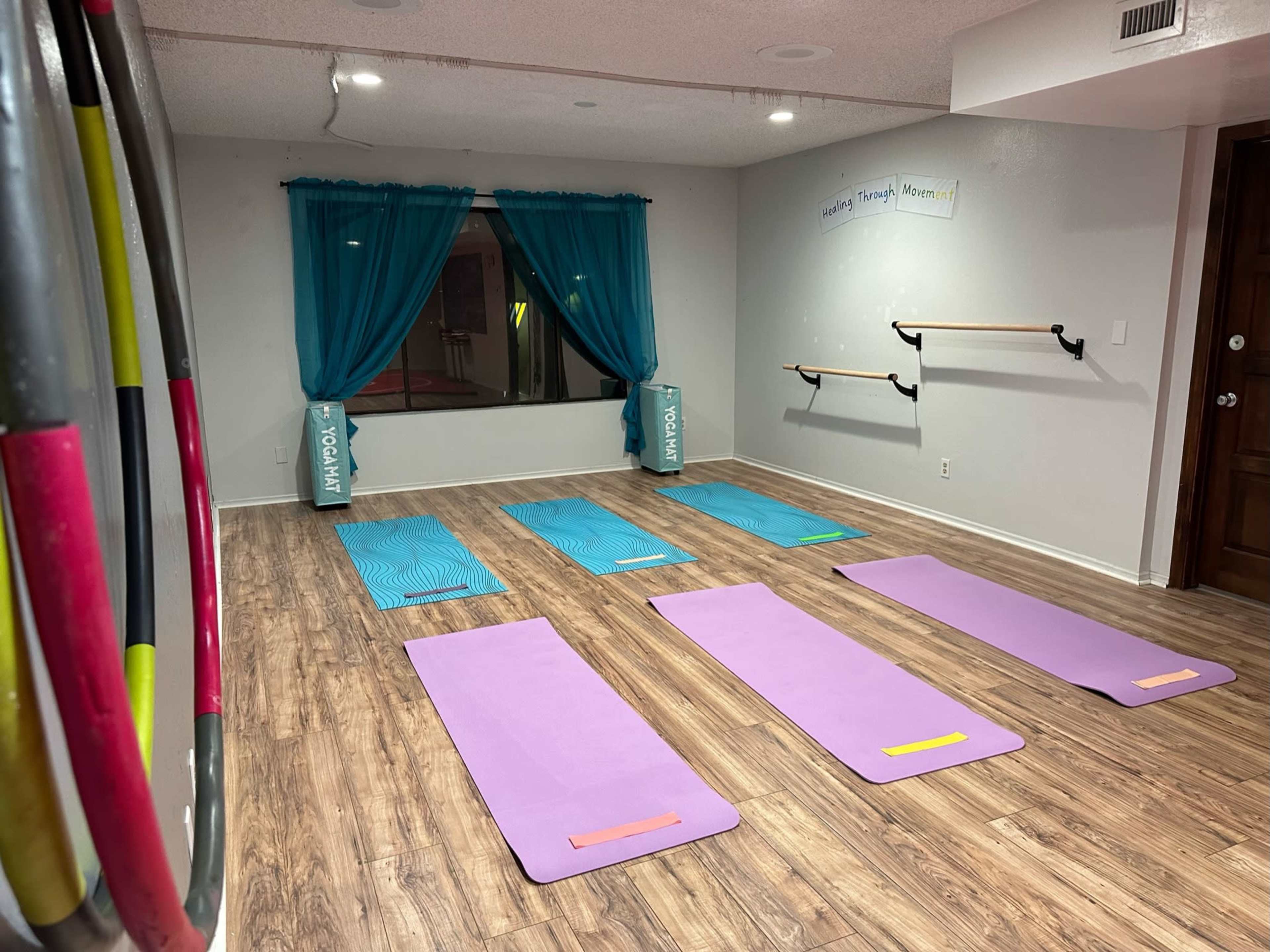 A fitness studio with wooden flooring, purple yoga mats arranged in rows, and turquoise curtains on the windows.