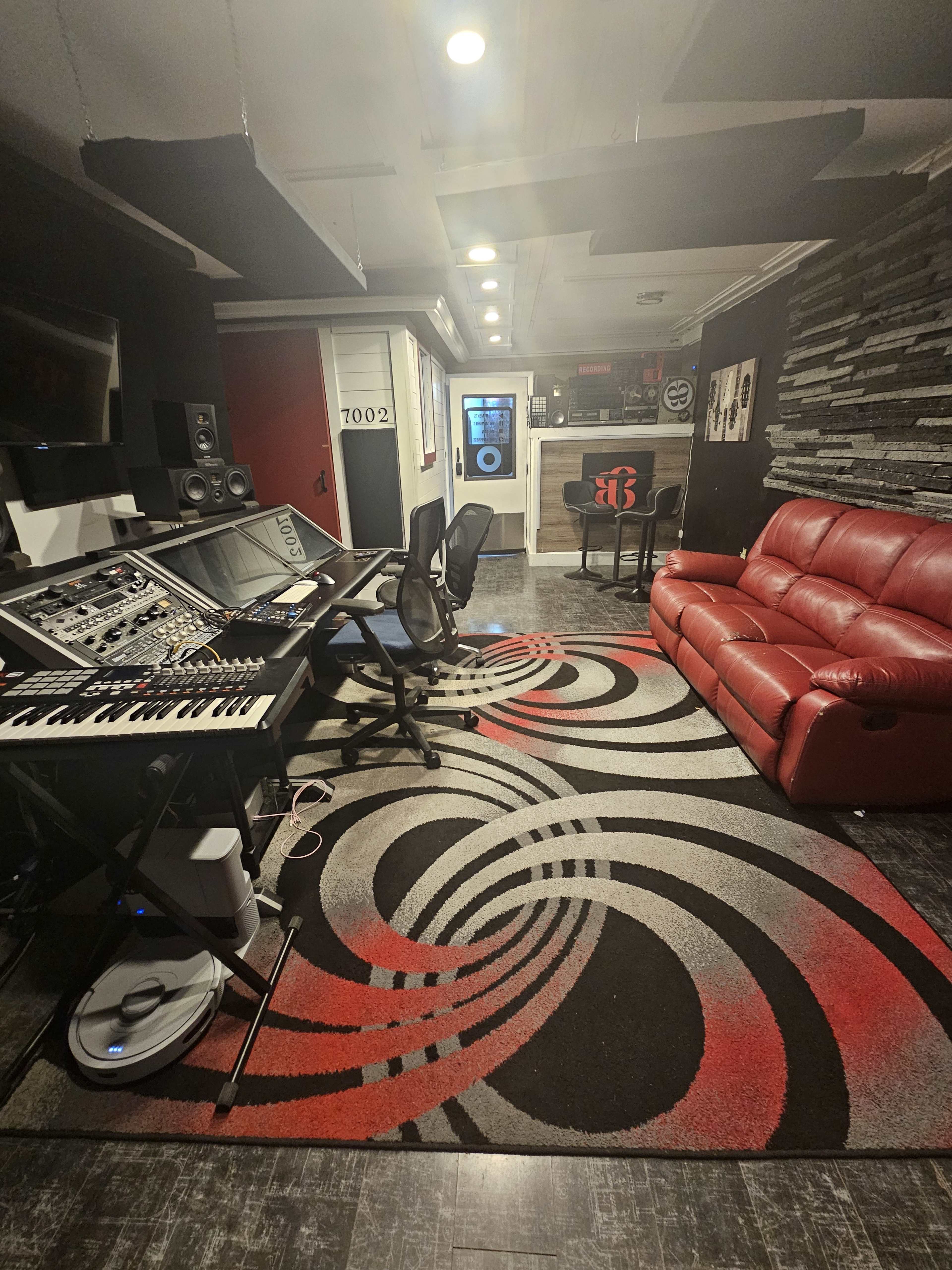 The image shows a music production studio featuring a control desk with audio equipment, a black chair, a red sofa, and a patterned rug on the floor.