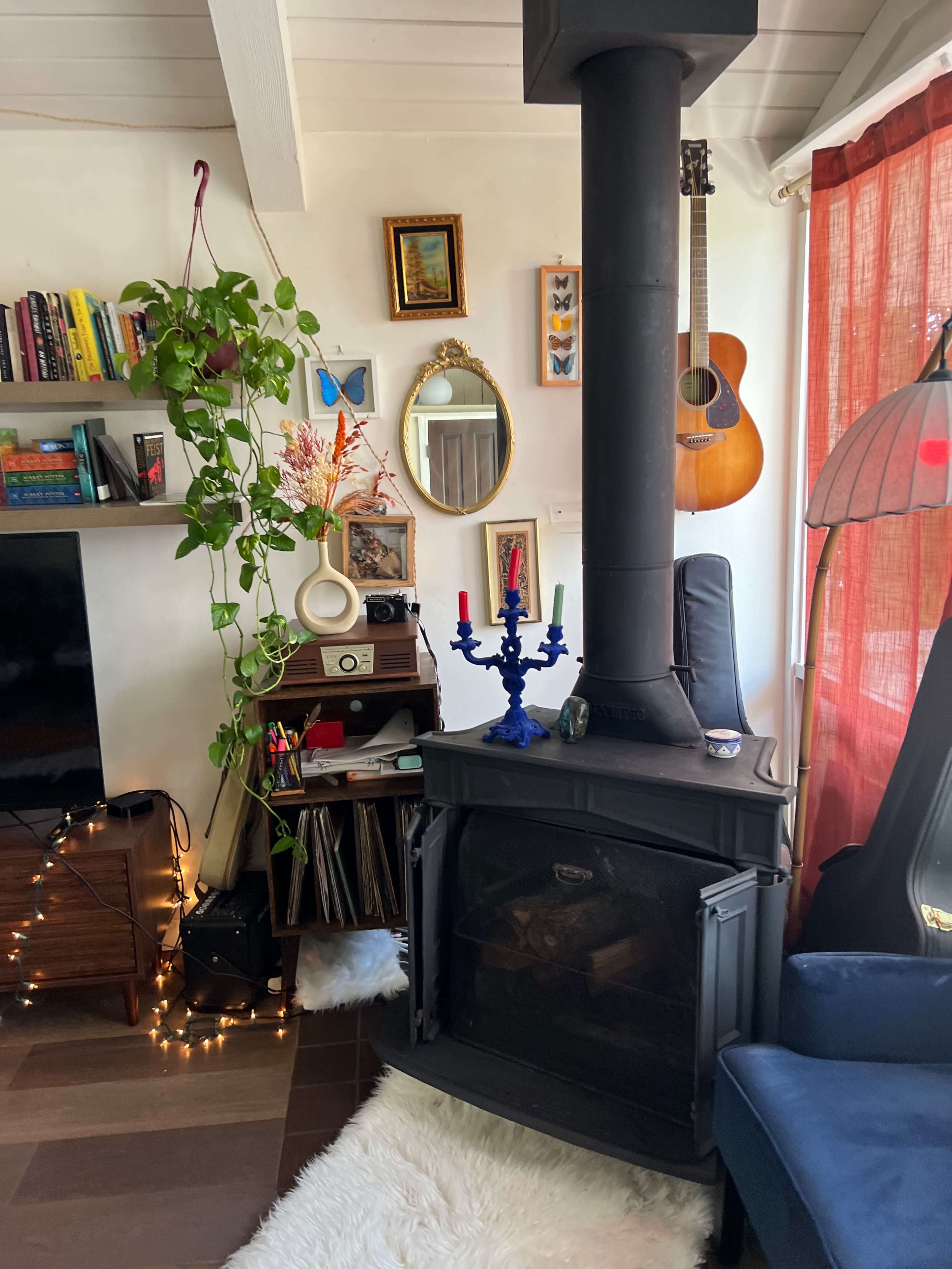 The image shows a cozy living room featuring a black wood stove, a guitar hanging on the wall, various books on a shelf, a decorative mirror, and houseplants, with warm lighting from a lamp and fairy lights.