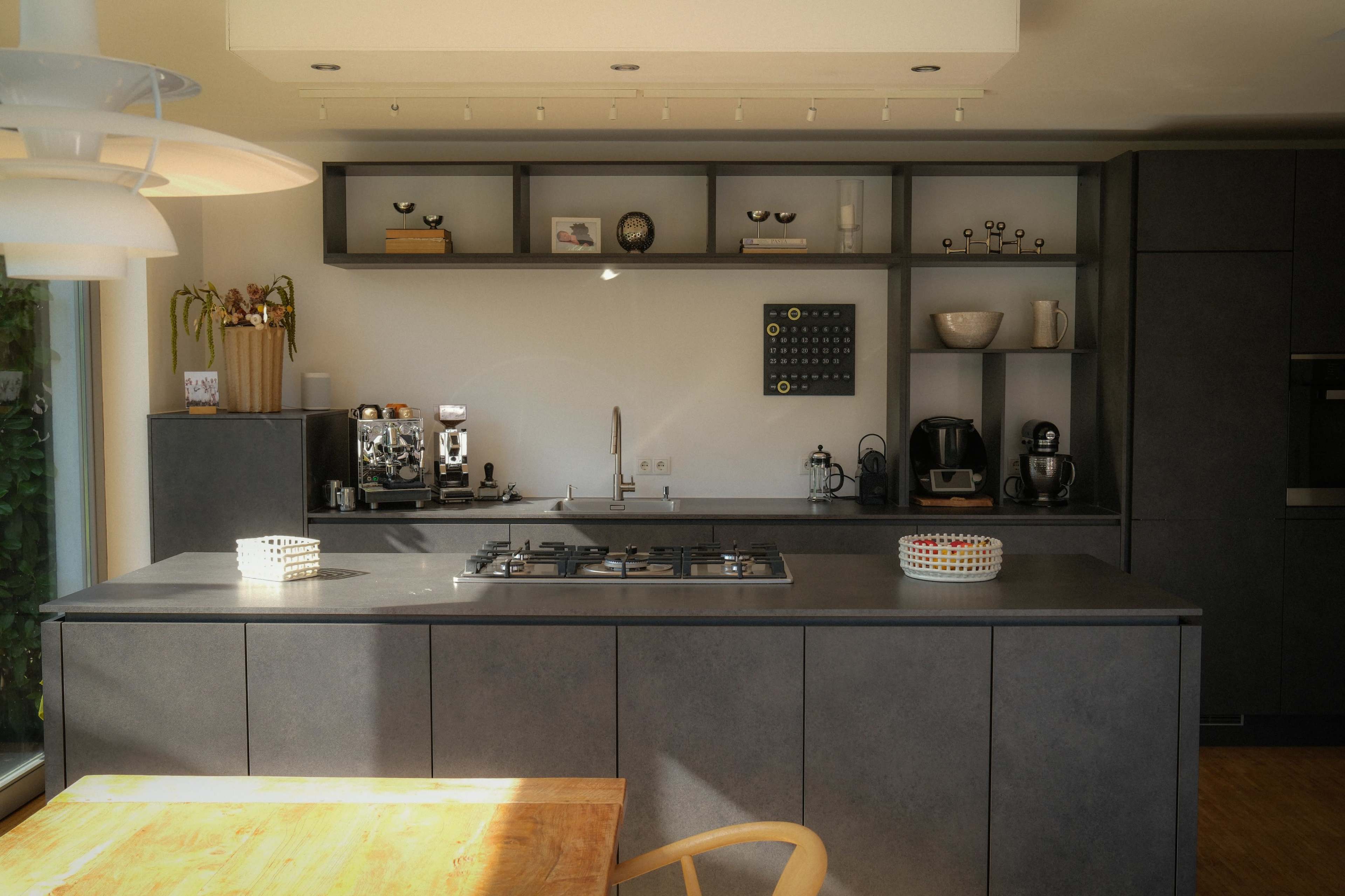 The image shows a modern kitchen with dark cabinetry, a countertop stove, and various kitchen appliances arranged on shelves and countertops.