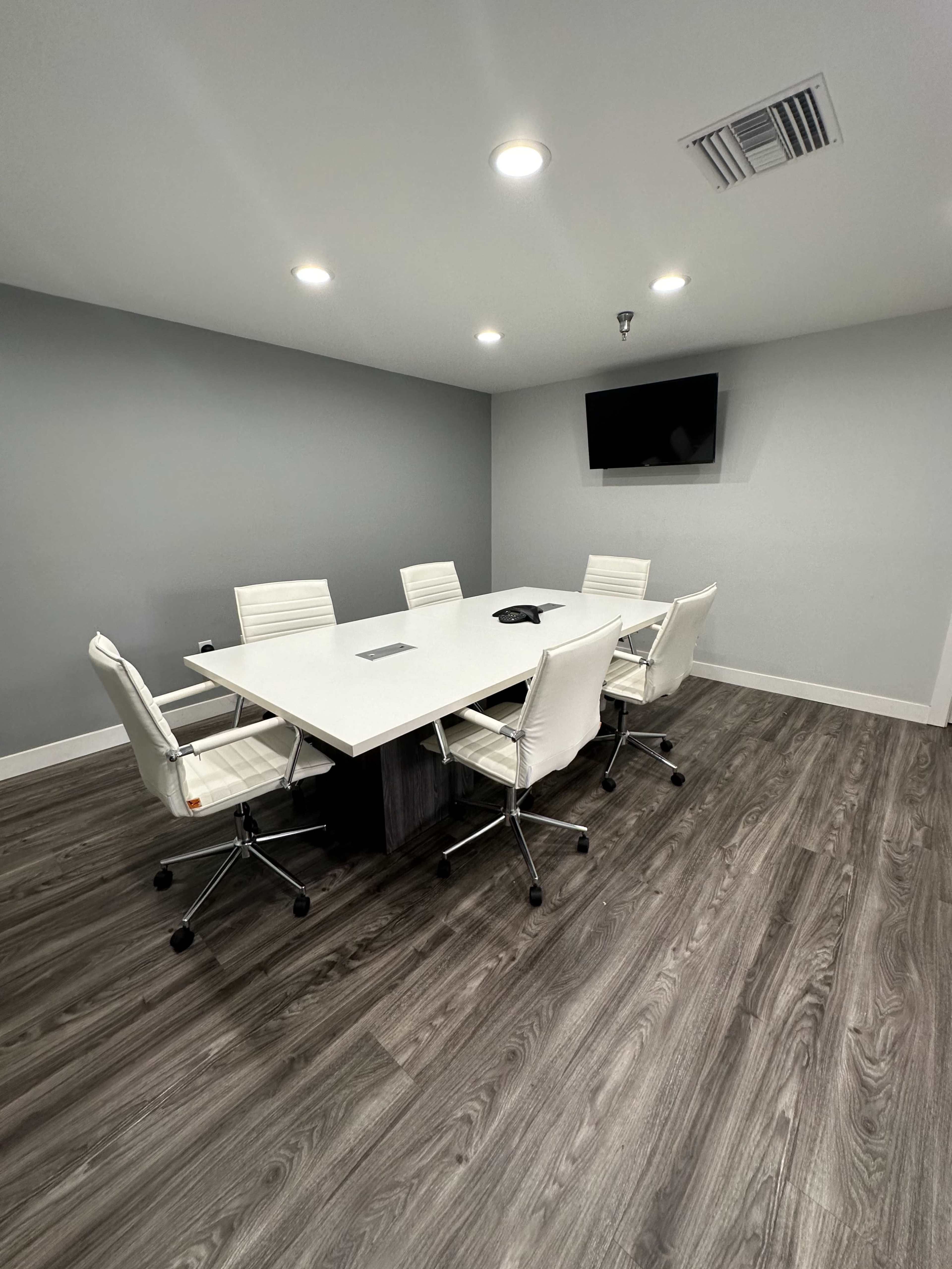 A modern conference room features a large white table surrounded by six ergonomic chairs, with a wall-mounted television and gray walls.