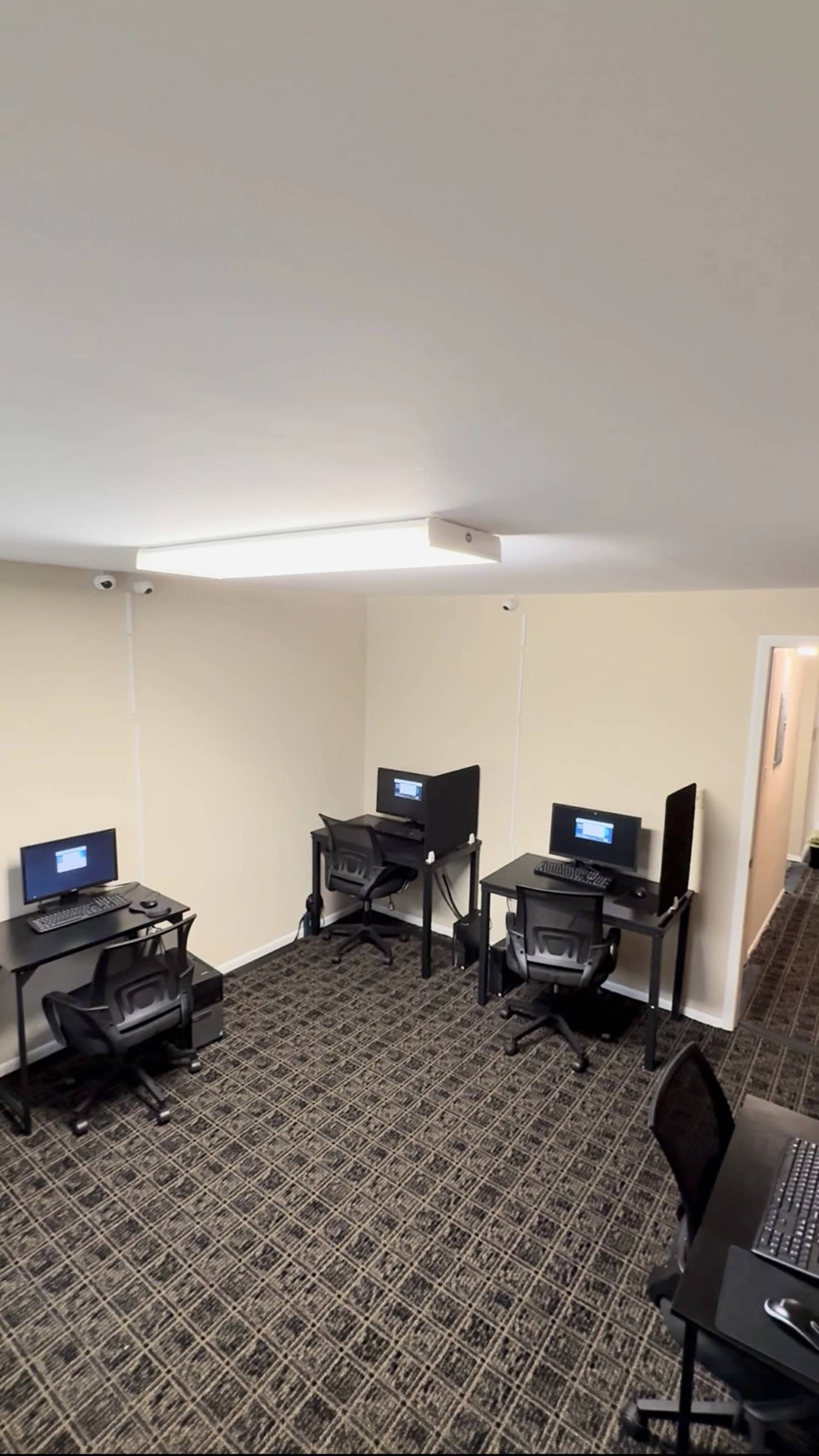 The image shows a small room with four computer workstations arranged against the walls, each featuring a monitor and an office chair, along with a carpeted floor and neutral-colored walls.