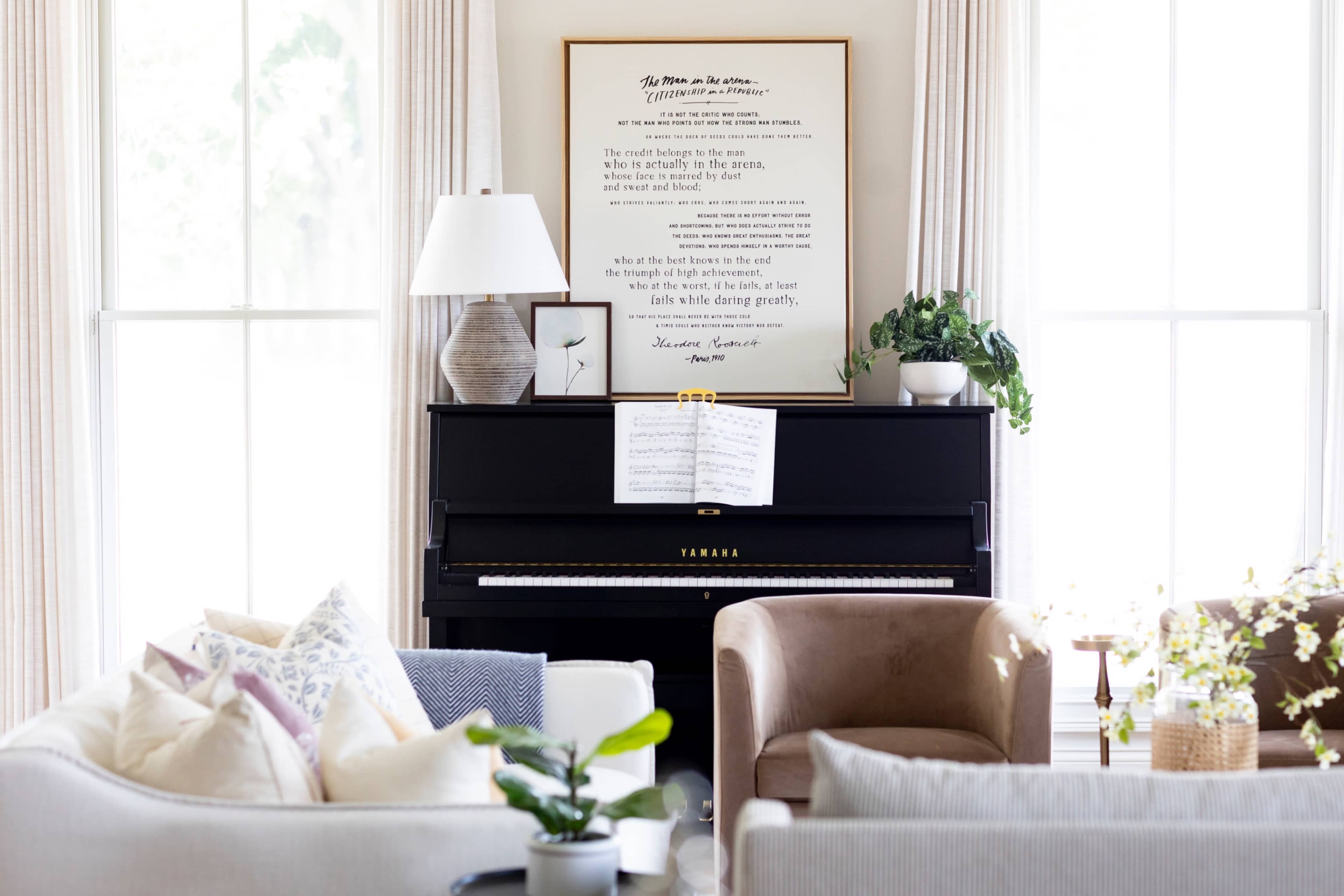 A black piano is placed against a wall adorned with a framed poem, surrounded by light-colored furniture and decorative plants in a bright living room.