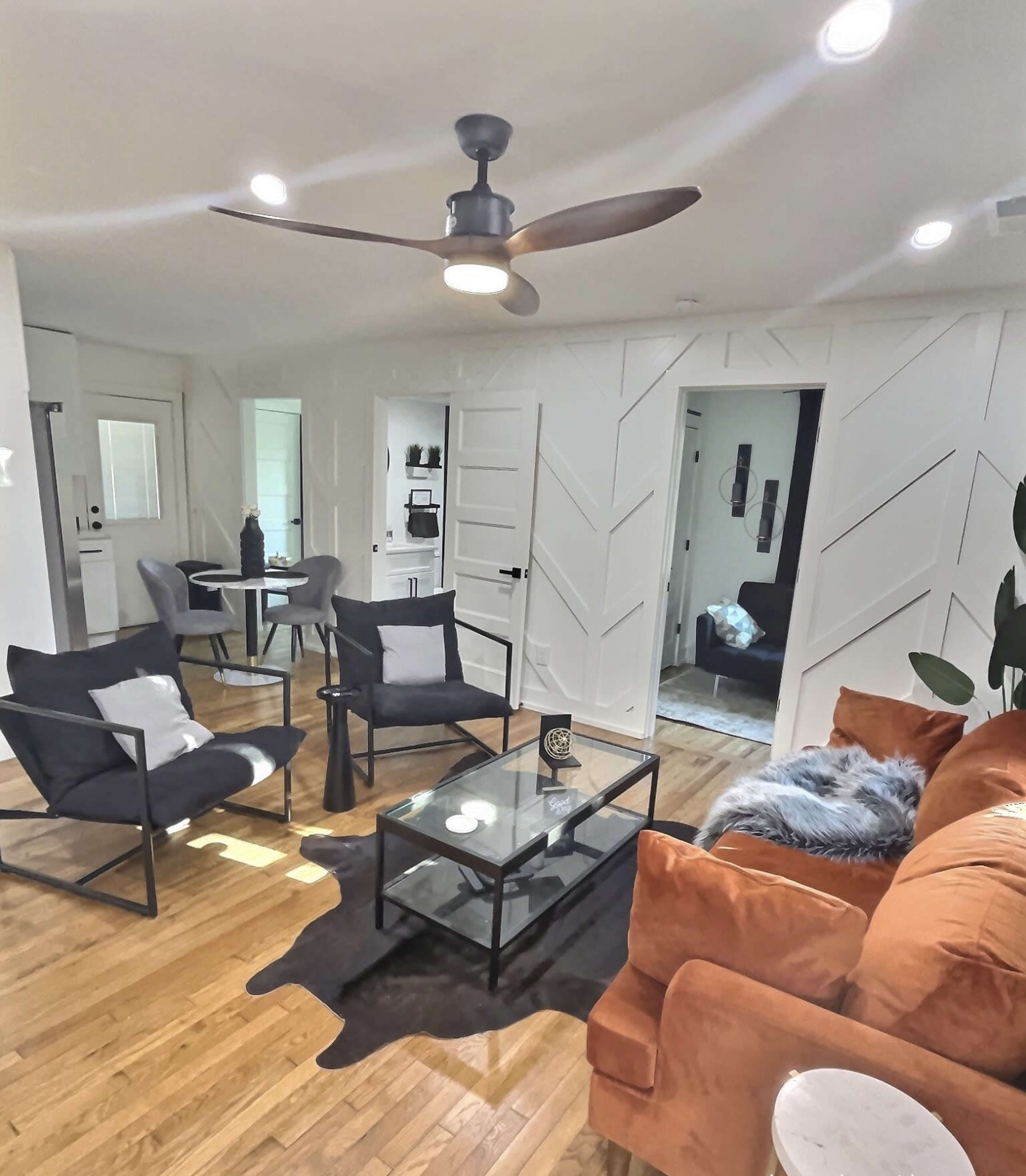 A modern living room features a brown sofa, black chairs, a glass coffee table on a cowhide rug, and a ceiling fan.