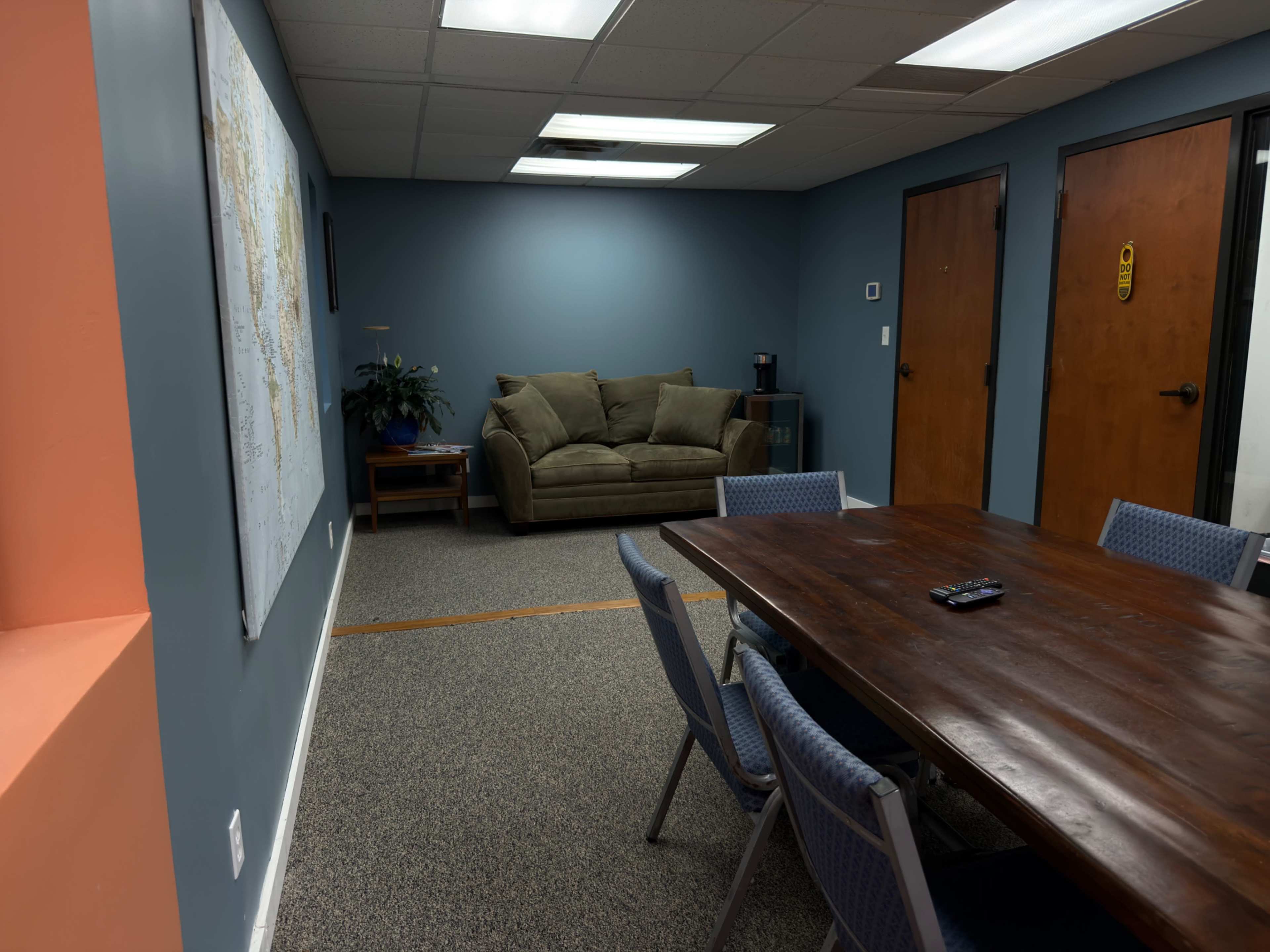 The image shows a conference room featuring a wooden table surrounded by chairs, a couch in the corner, and a map on the wall.