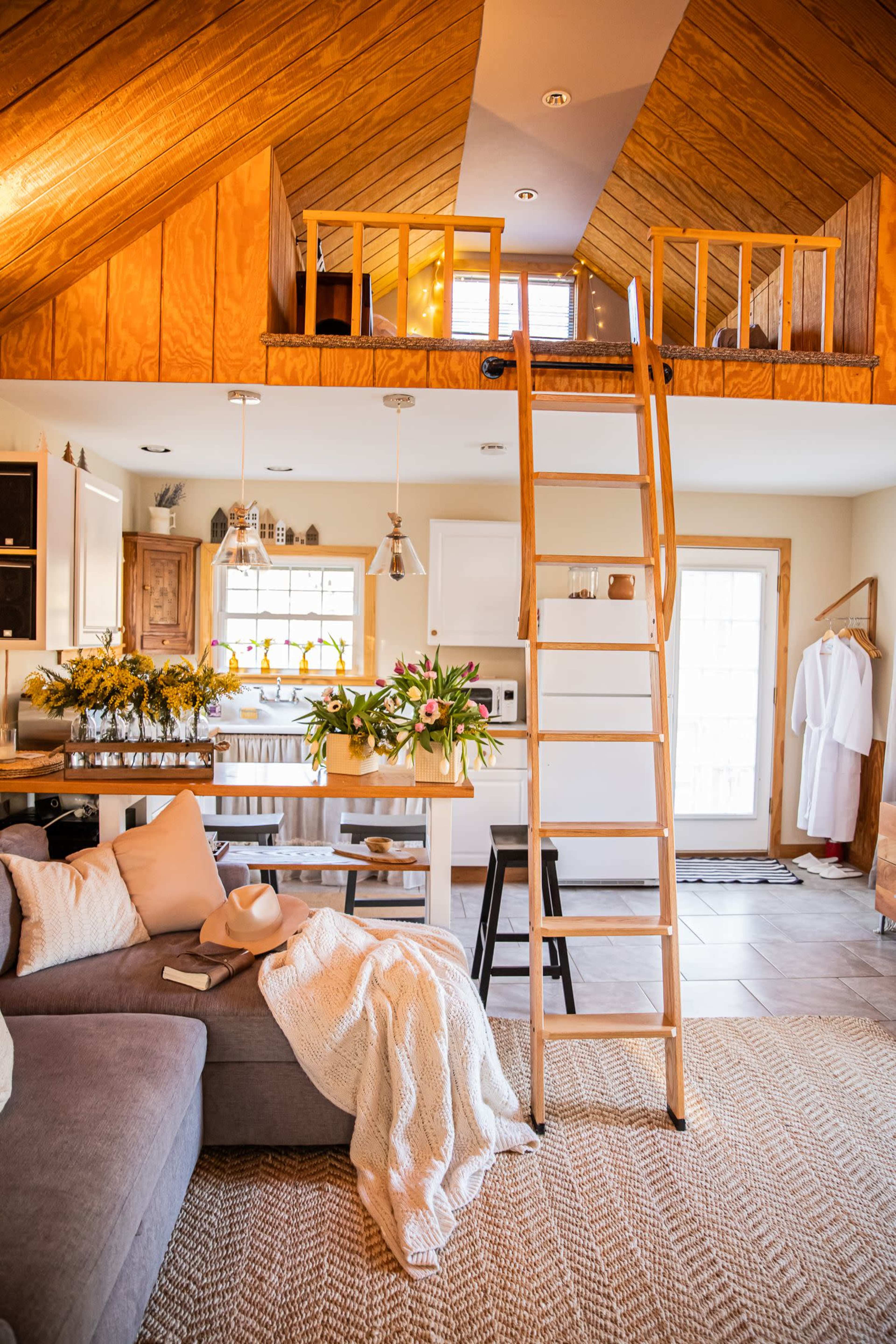 A cozy interior of a small cabin with a loft, featuring a ladder, a sectional sofa, and a kitchen area, adorned with flowers and warm lights.