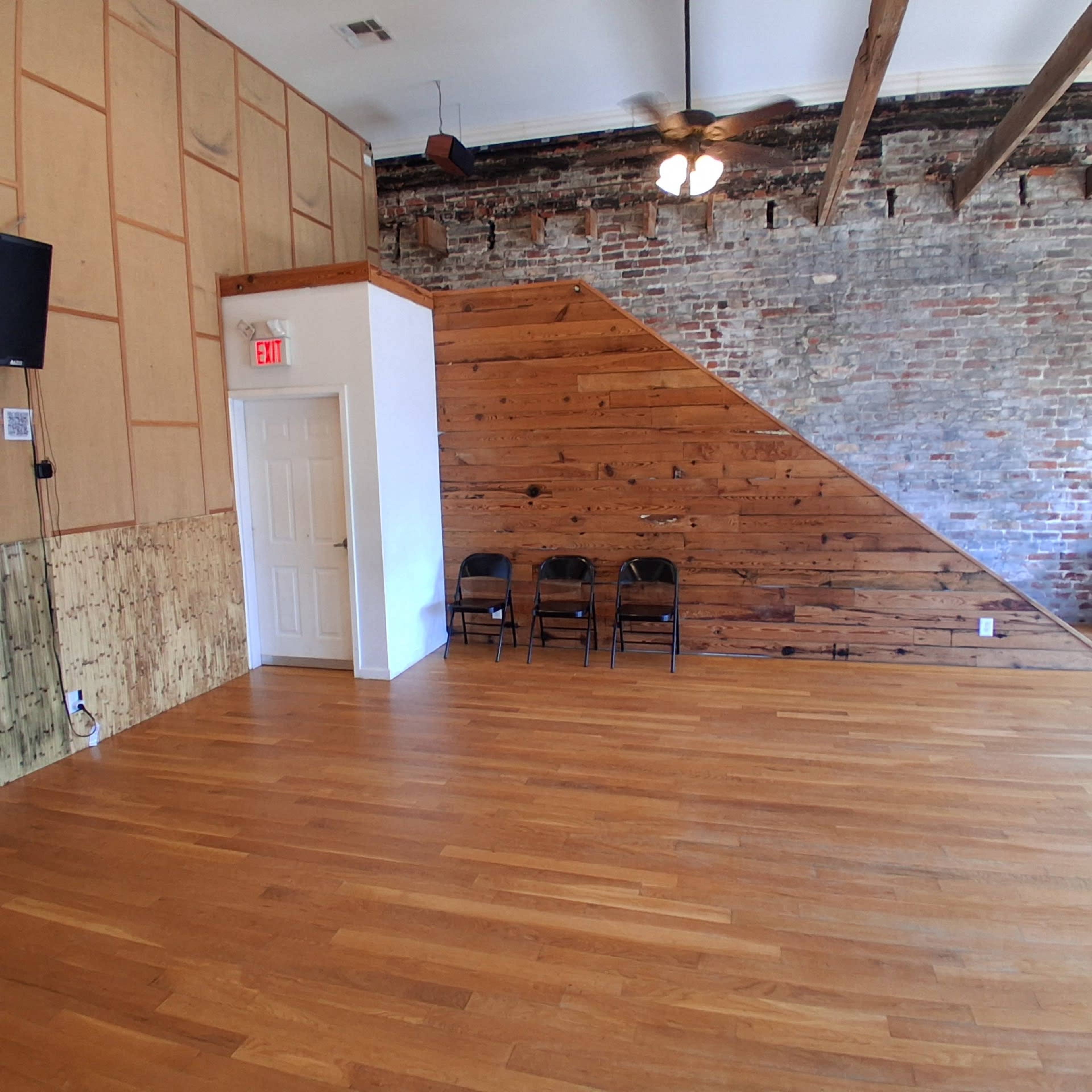The image shows an interior space with wooden flooring, exposed brick walls, and a section featuring wooden paneling, along with three black chairs arranged against one wall.