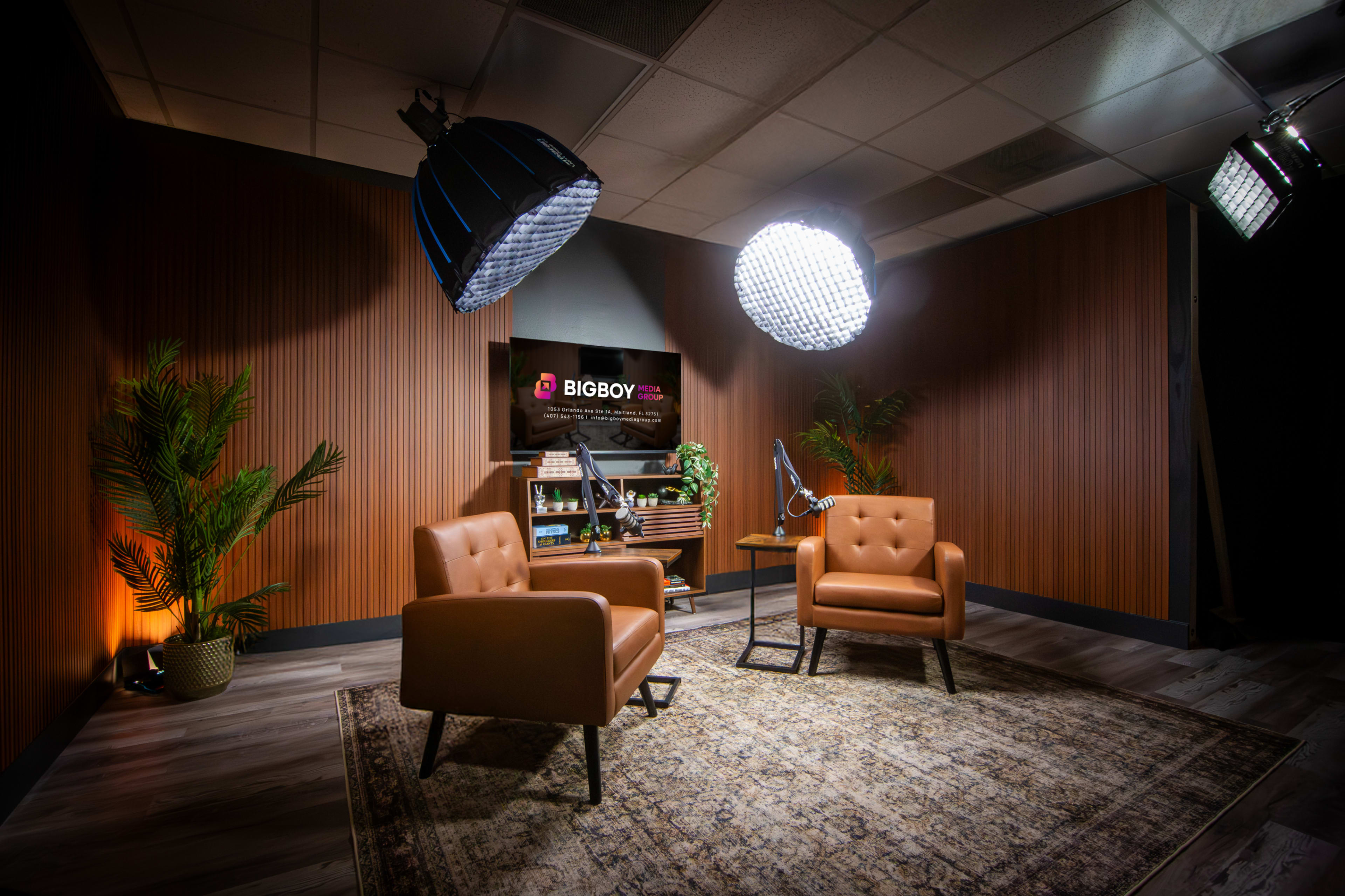 The image shows a well-lit studio setup with two brown leather chairs, a decorative rug, and a backdrop featuring a shelf and a sign that reads "BIGBOY."