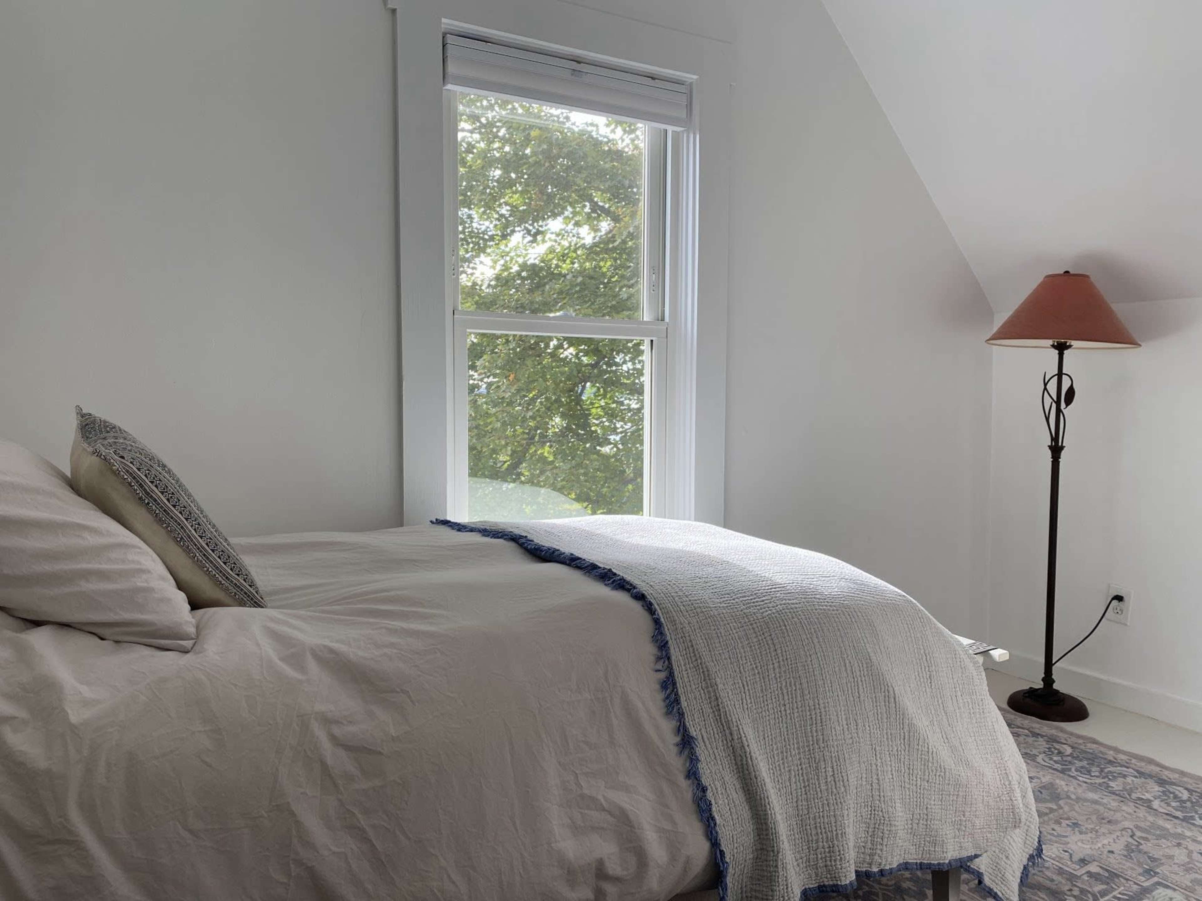 A neatly made bed with a light-colored blanket sits in a bright bedroom featuring a window and a floor lamp.