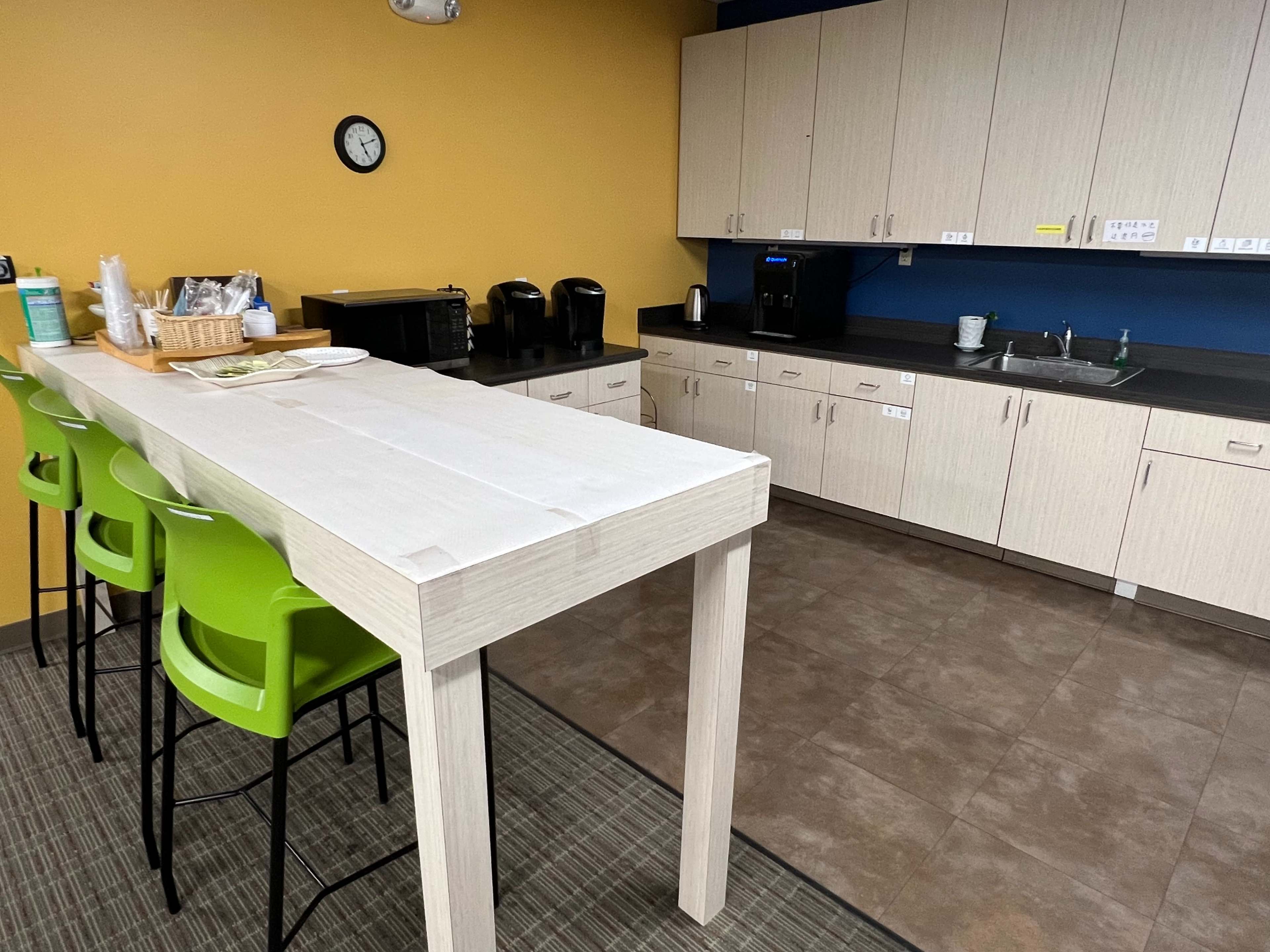 A kitchen area with a long white table and four green stools, featuring appliances such as coffee makers and a sink along the counter.