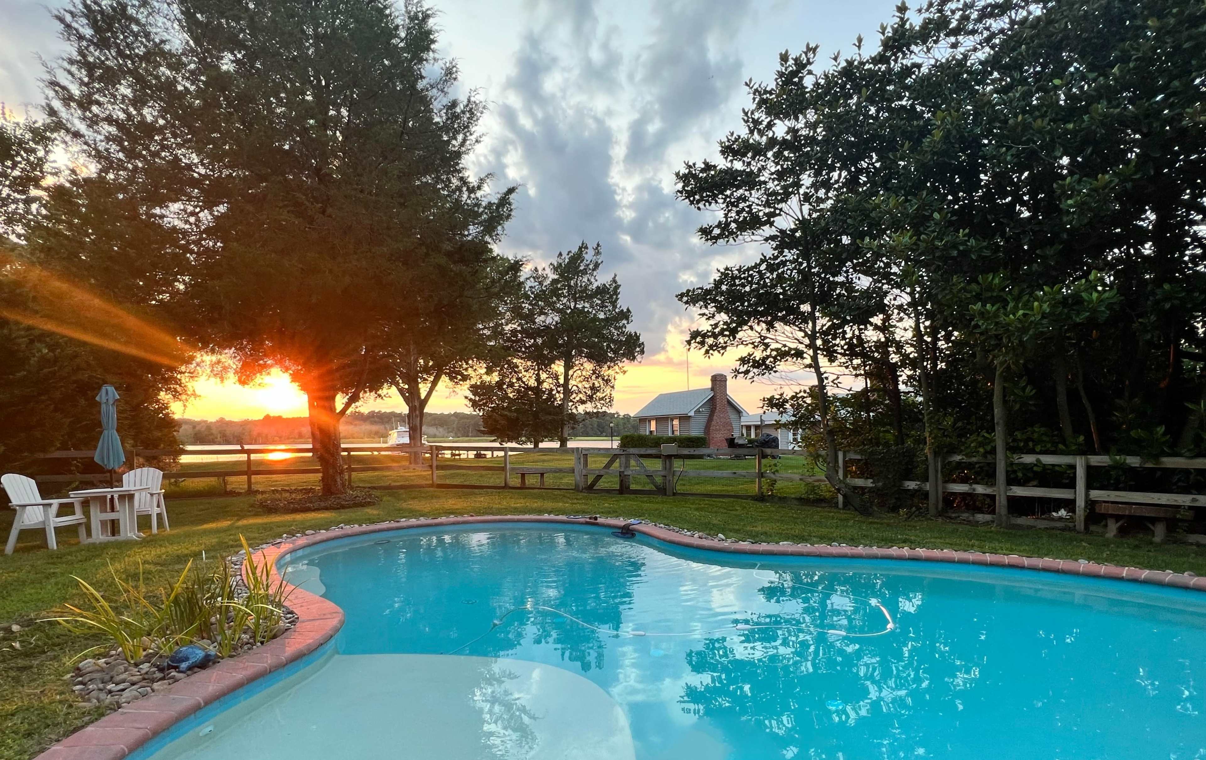 A tranquil backyard scene with a swimming pool reflecting the sunset and a nearby house surrounded by trees.