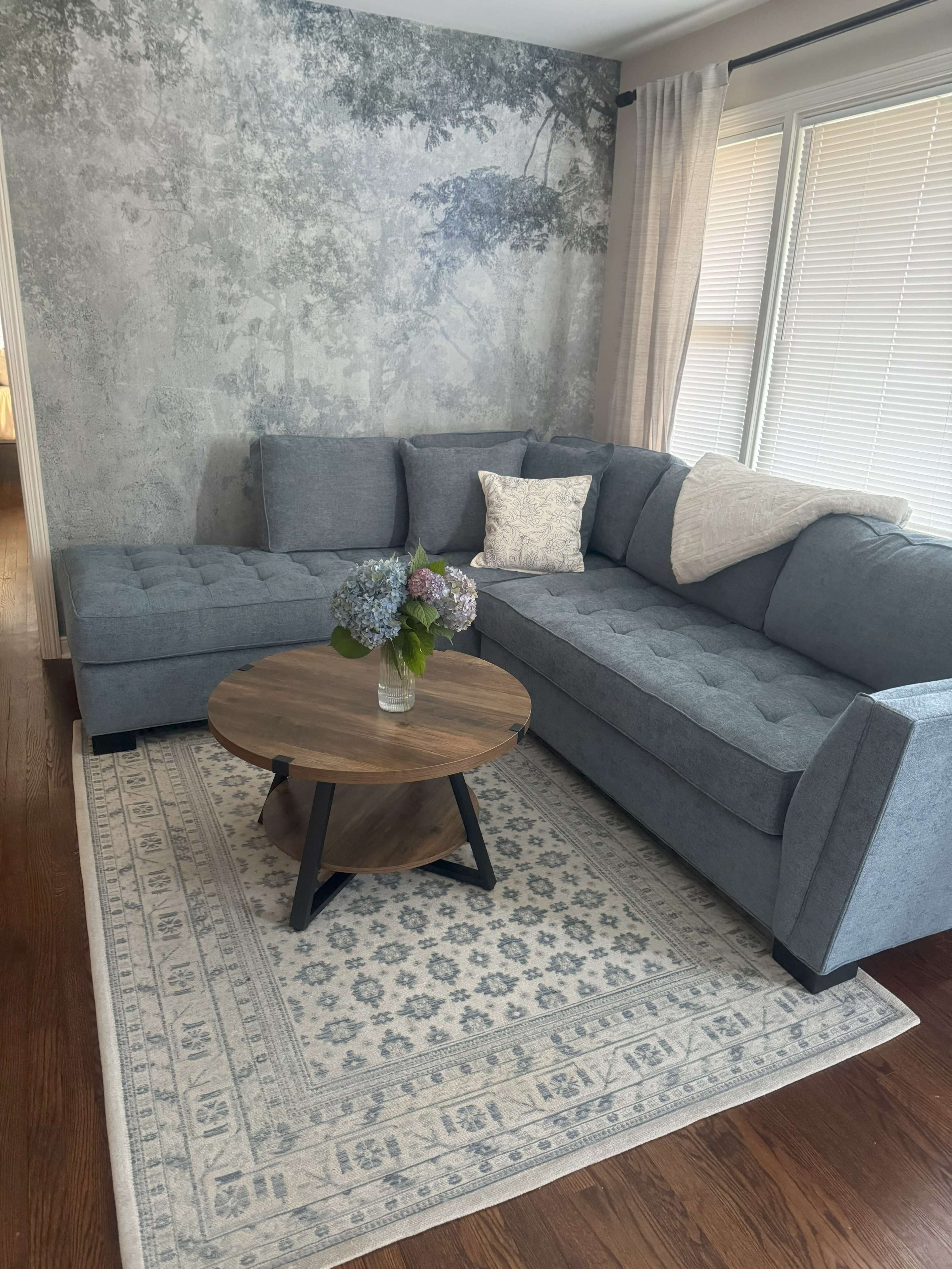 A living room corner with a gray sectional sofa, a round wooden coffee table, and a floral arrangement on top of a patterned area rug, against a wall featuring a mural.