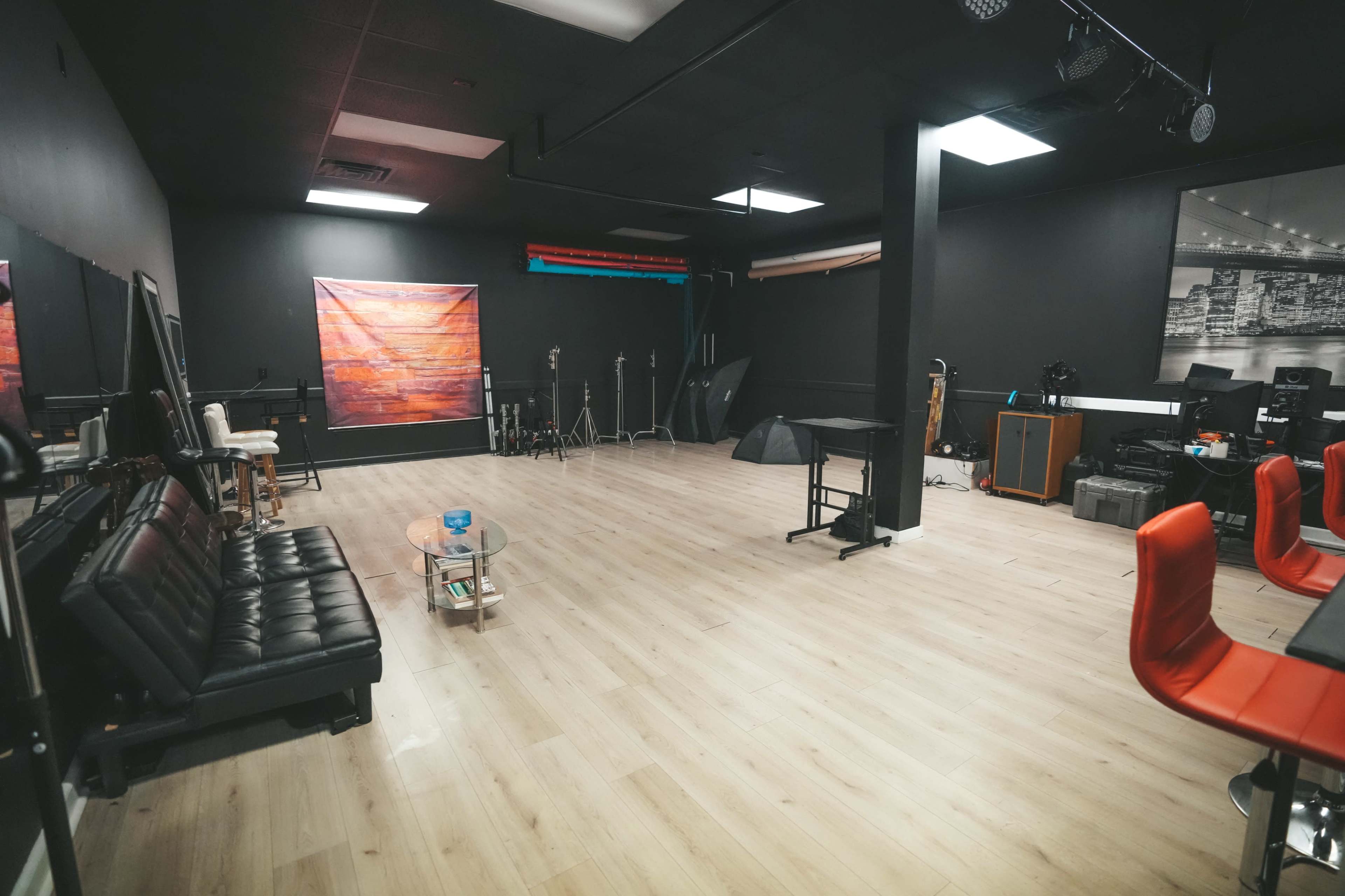 The image shows a spacious, dimly lit studio with wooden flooring, black walls, and various equipment laid out, including furniture and photography gear.