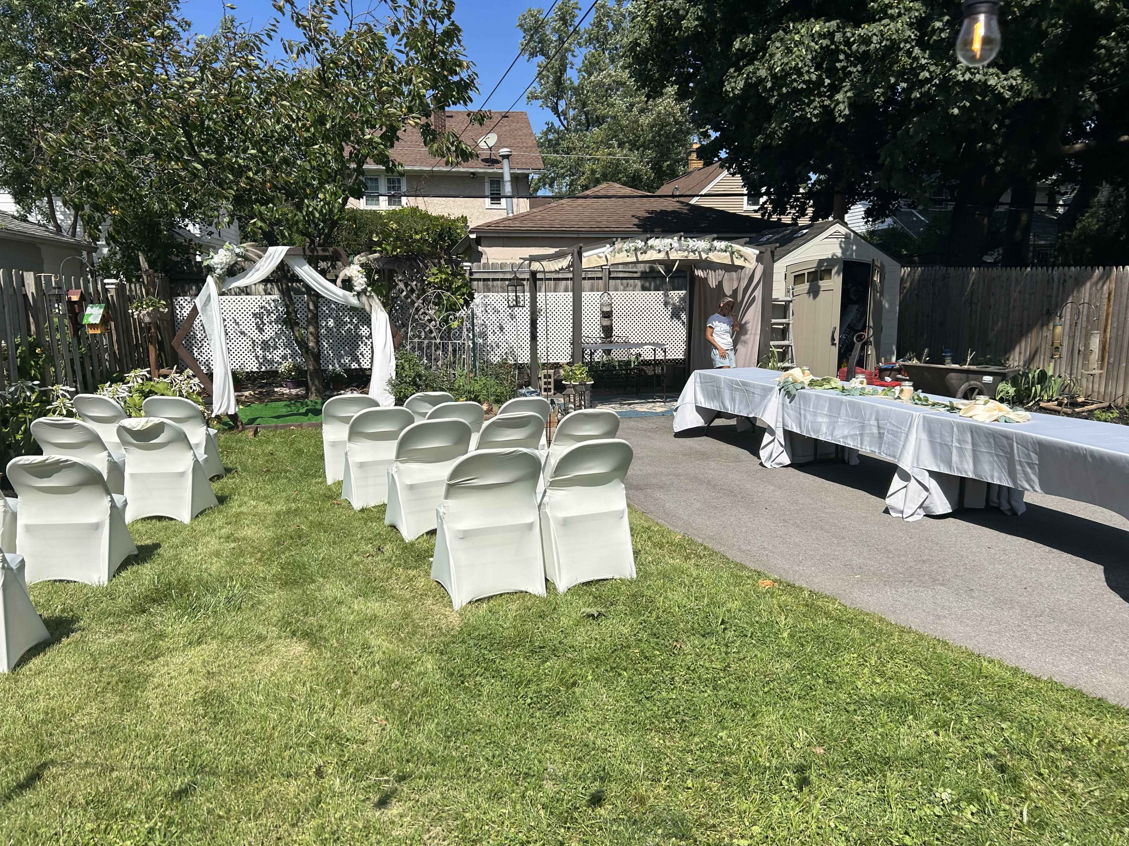 A neatly arranged outdoor event space features white chairs facing an arch decorated for a ceremony, with a long table set for a reception nearby.