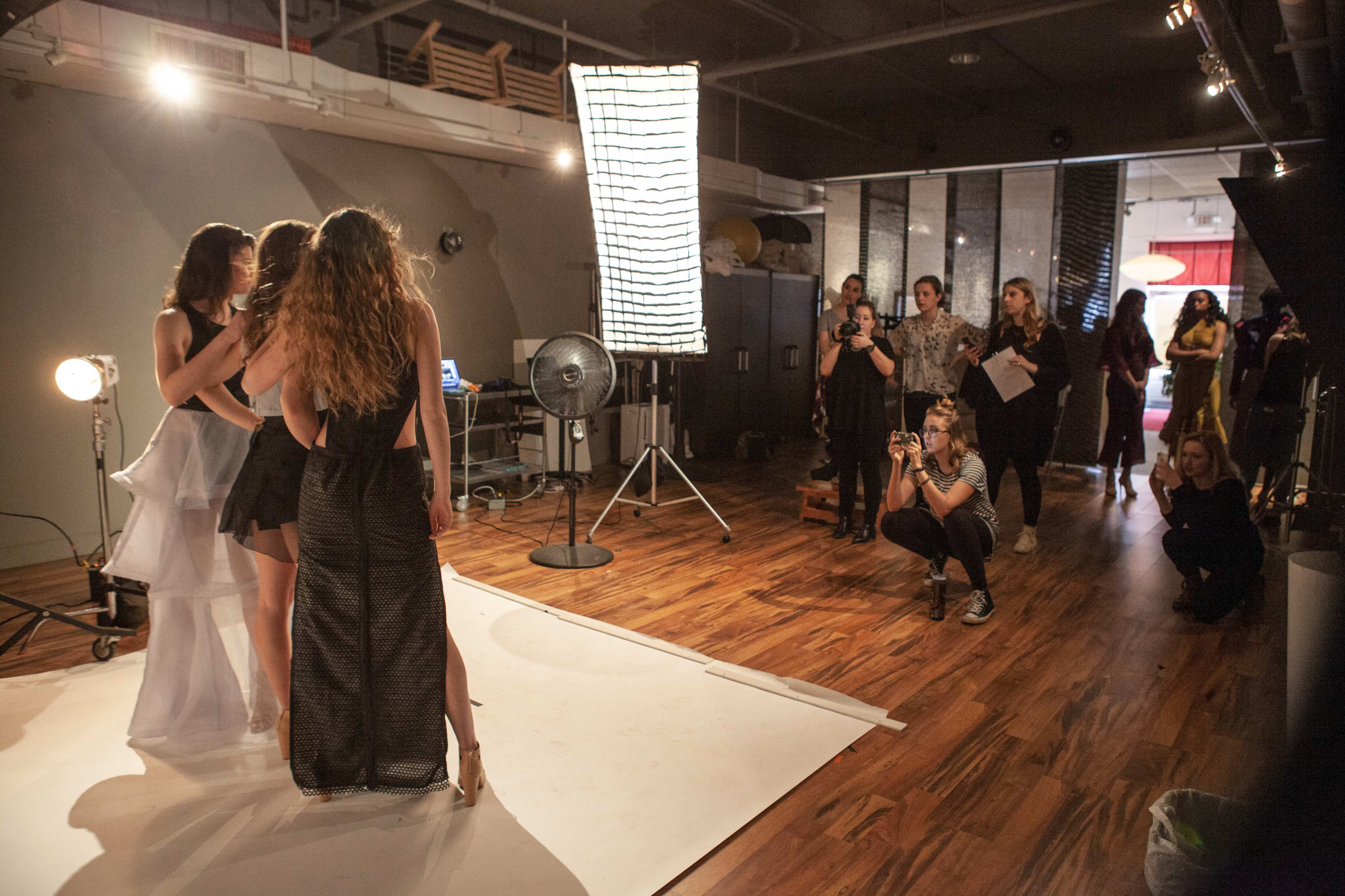 A group of models poses for a photoshoot while a photographer captures the scene in a studio with various lighting equipment and onlookers.