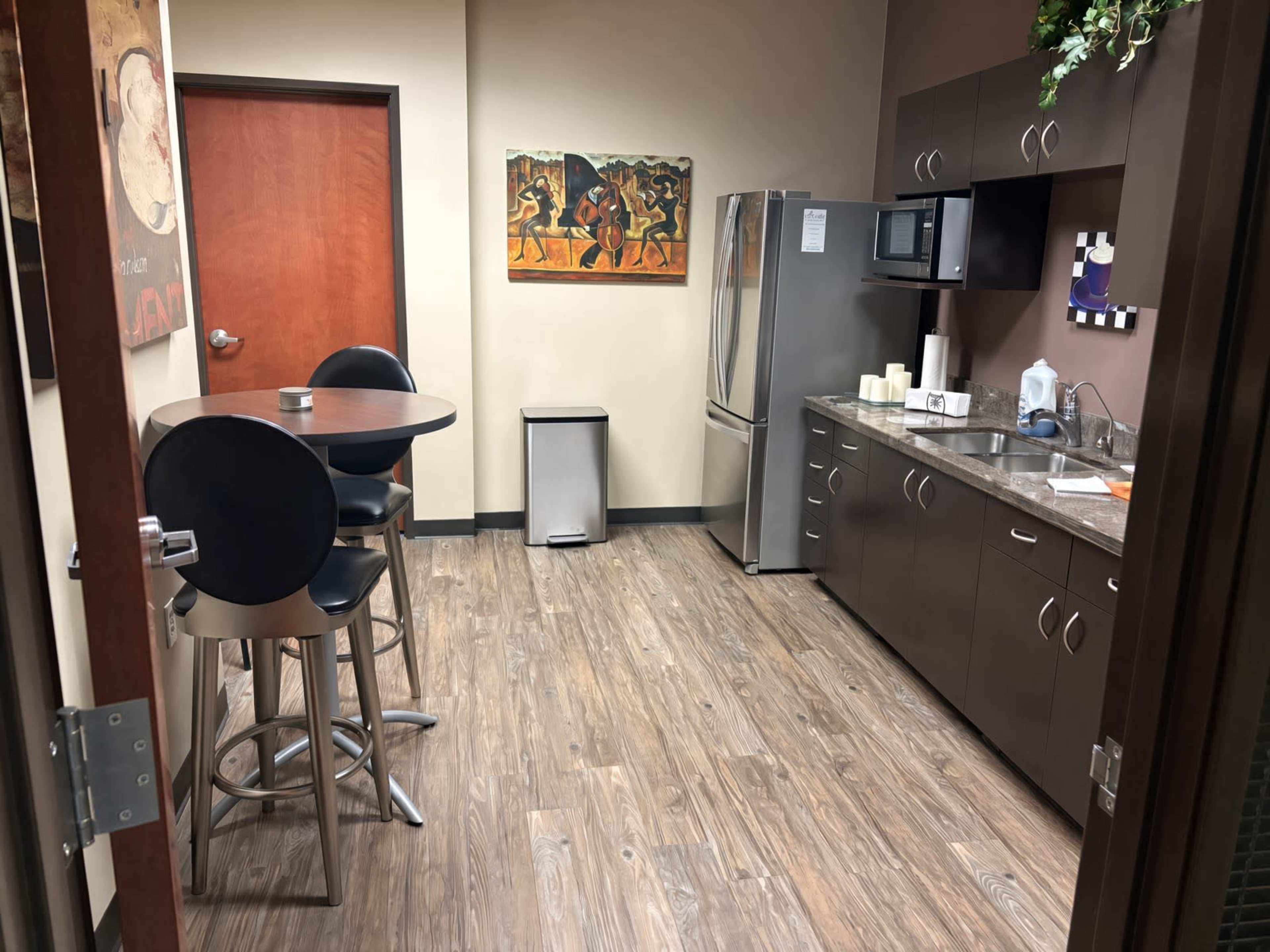 The image shows a break room with a small round table, two high stools, a refrigerator, a microwave, and countertop space, all set against a backdrop of wood-like flooring and painted walls with artwork.