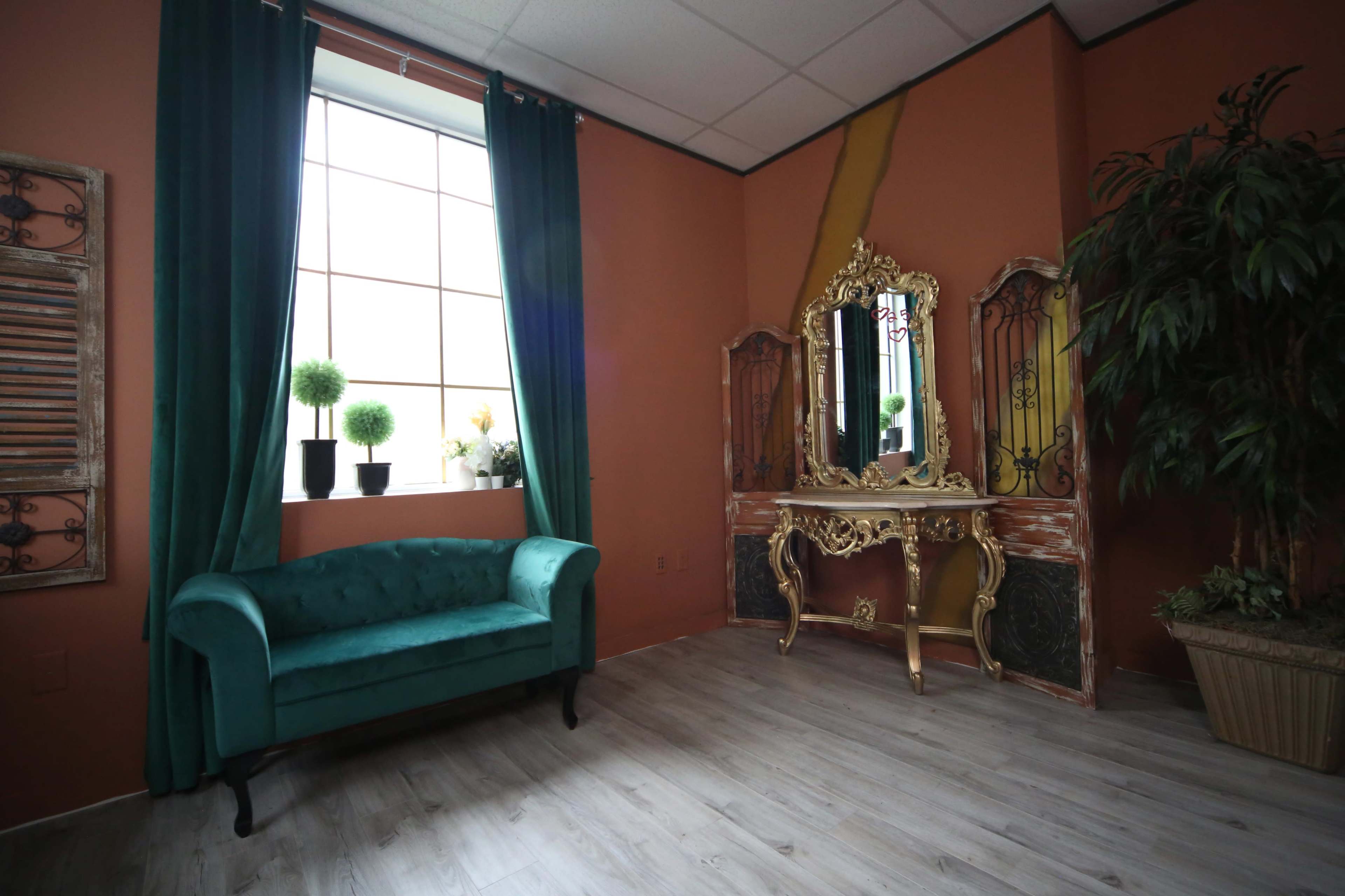 The room features a turquoise couch beside a large window with green curtains, a decorative gold mirror, and potted plants.