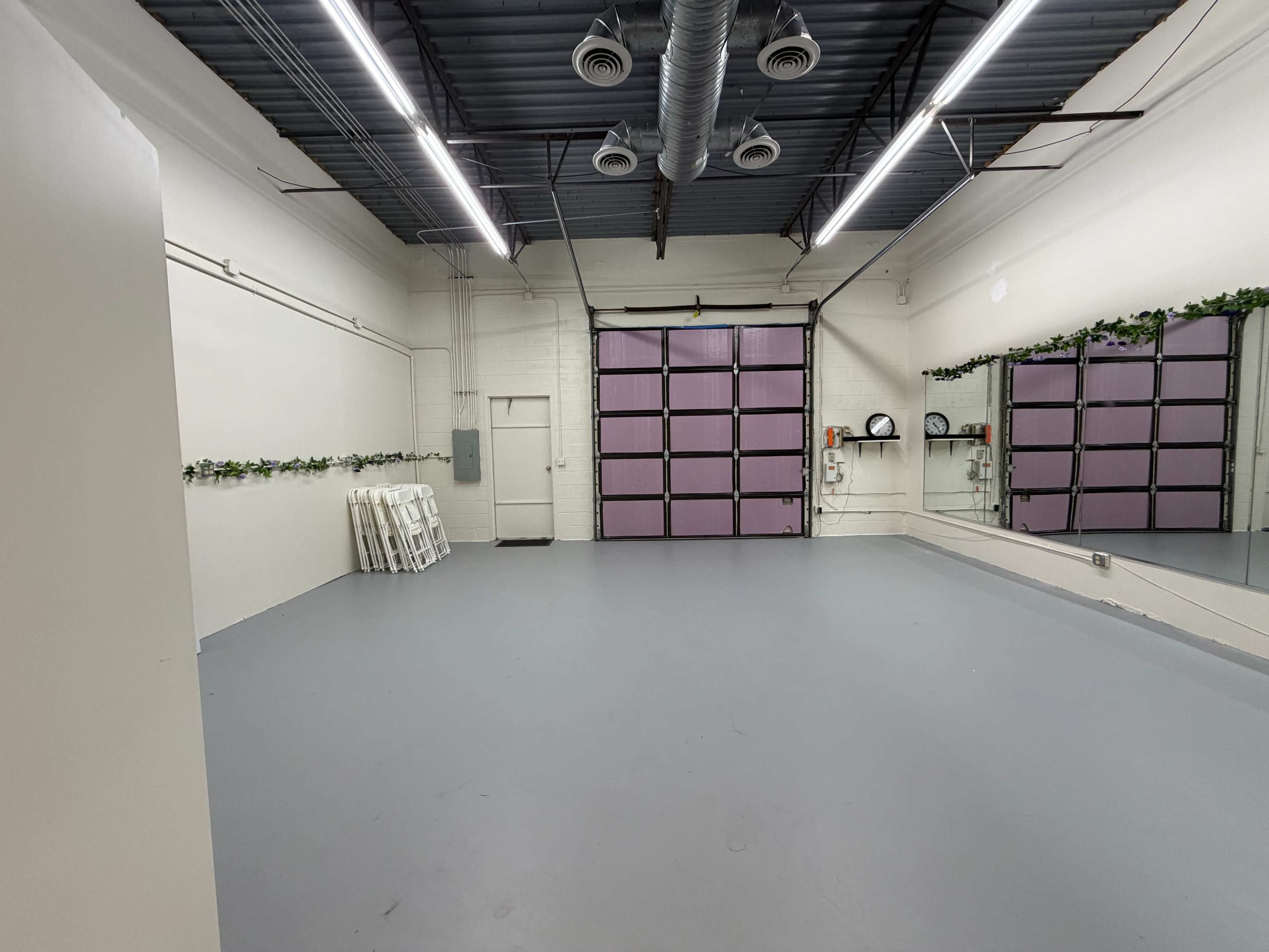 The image shows a spacious, empty studio with gray flooring, mirrored walls, and a large garage door with purple panels.