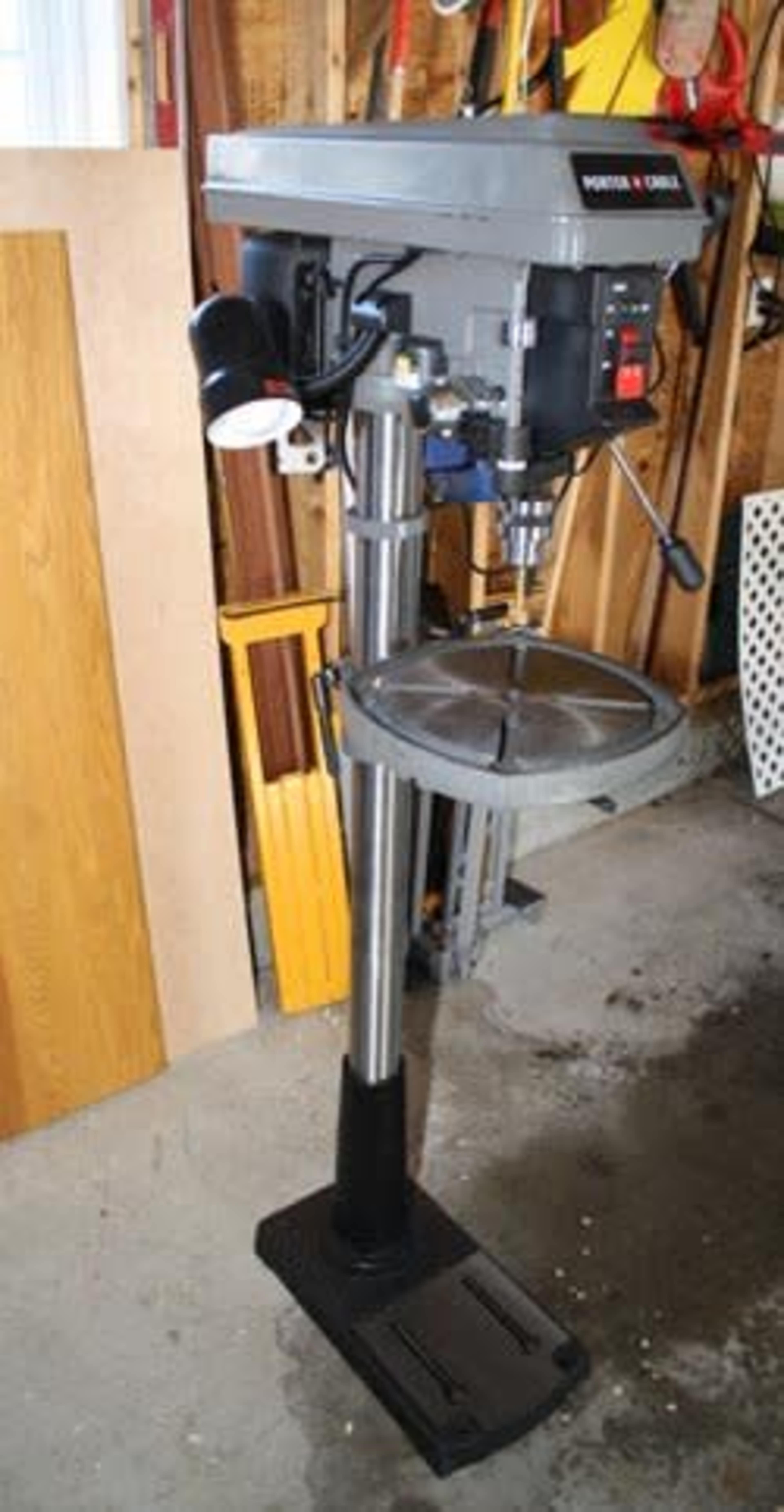 A gray drill press mounted on a black stand, positioned in a workshop with wooden panels and tools visible in the background.