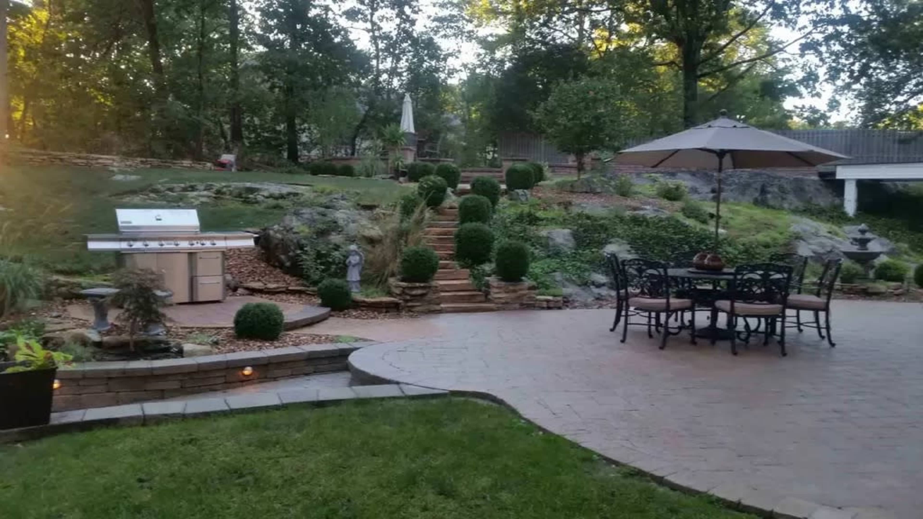 The image shows a landscaped backyard featuring a stone patio with a dining table and umbrella, a grill station, and stair steps leading to a garden area.
