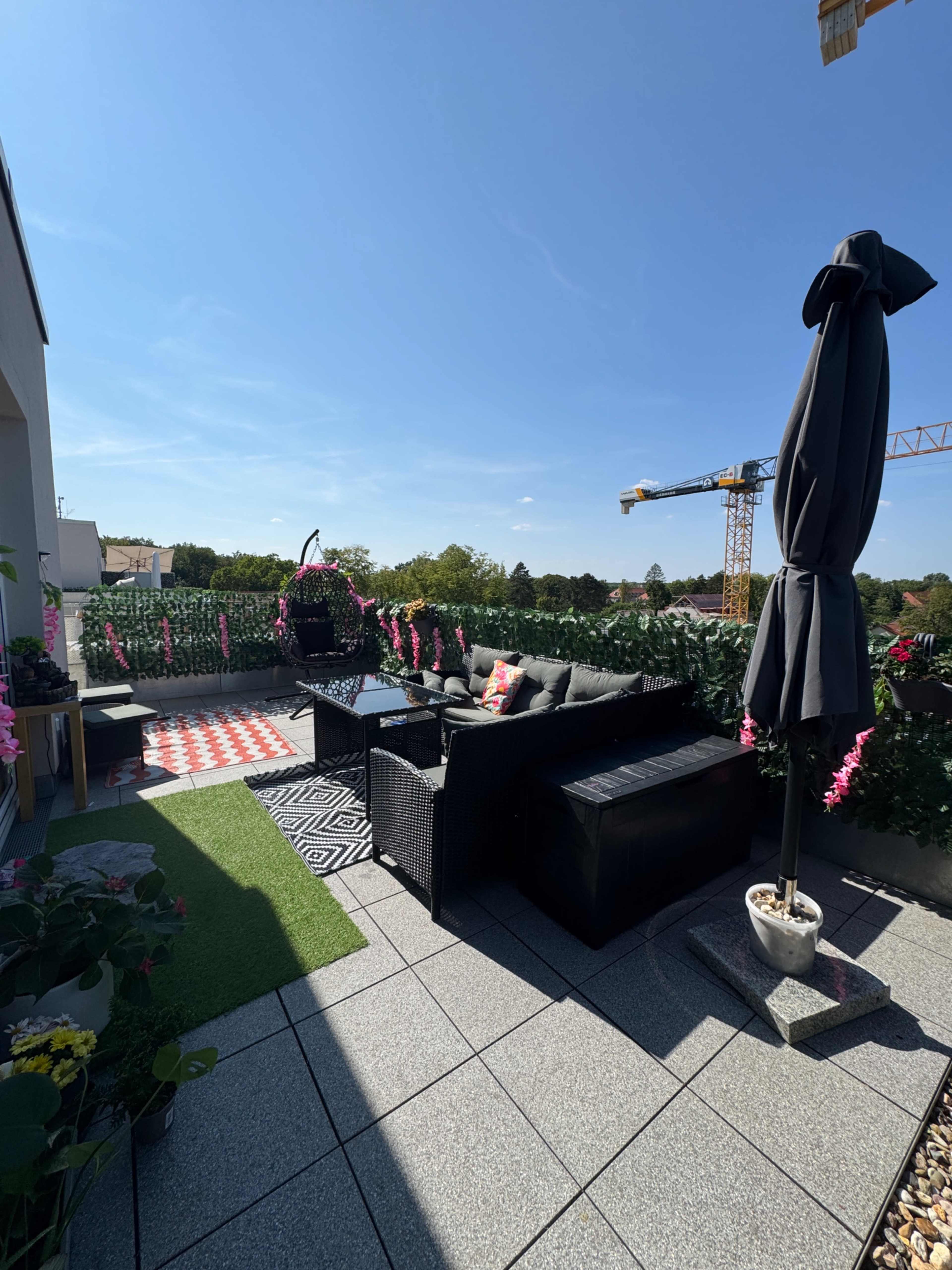 The image shows a balcony styled with outdoor furniture, a hanging chair, and vibrant flowers, under a clear blue sky.