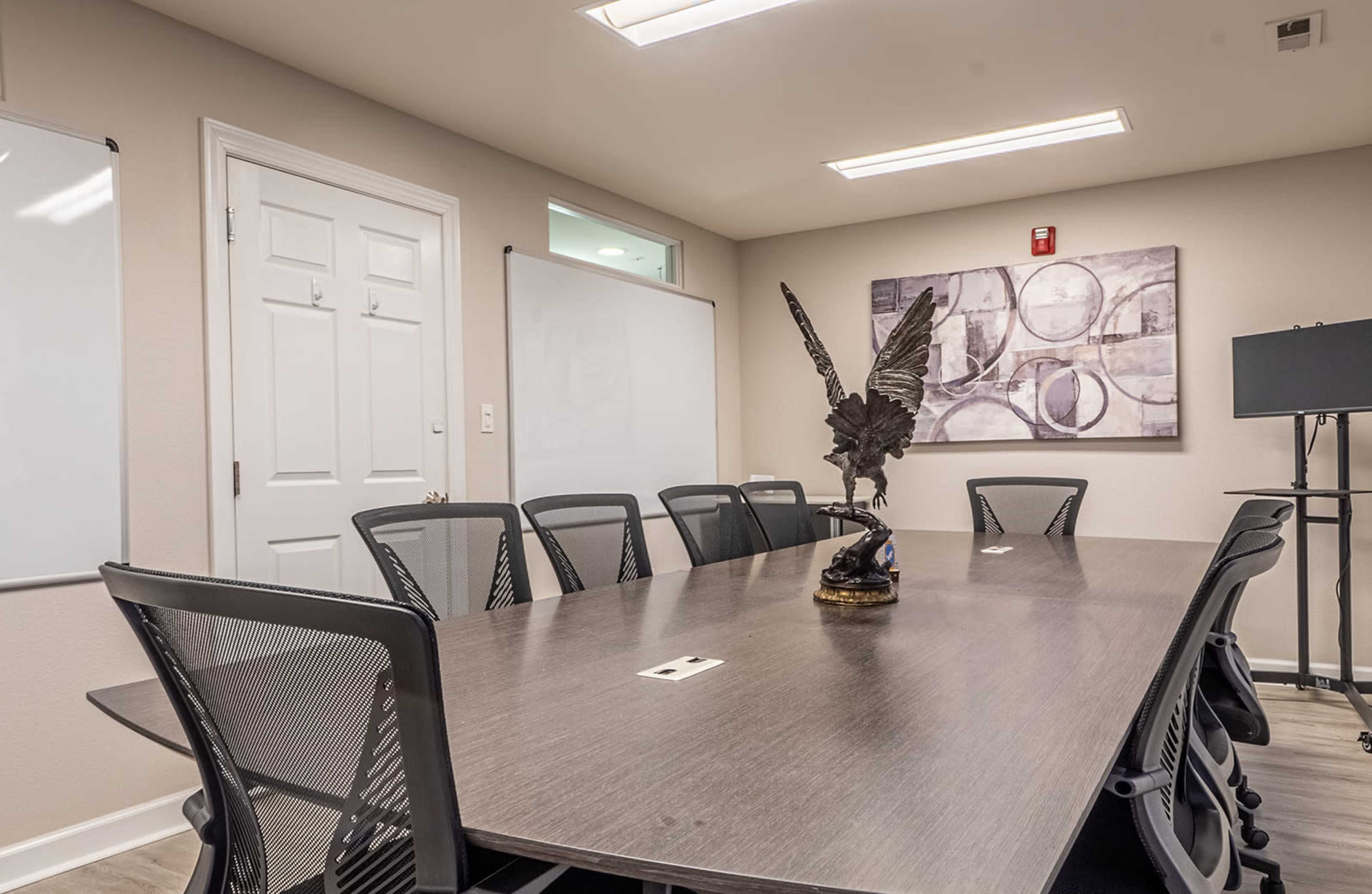 A conference room features a long table surrounded by black mesh chairs, with a decorative sculpture and whiteboards on the walls.