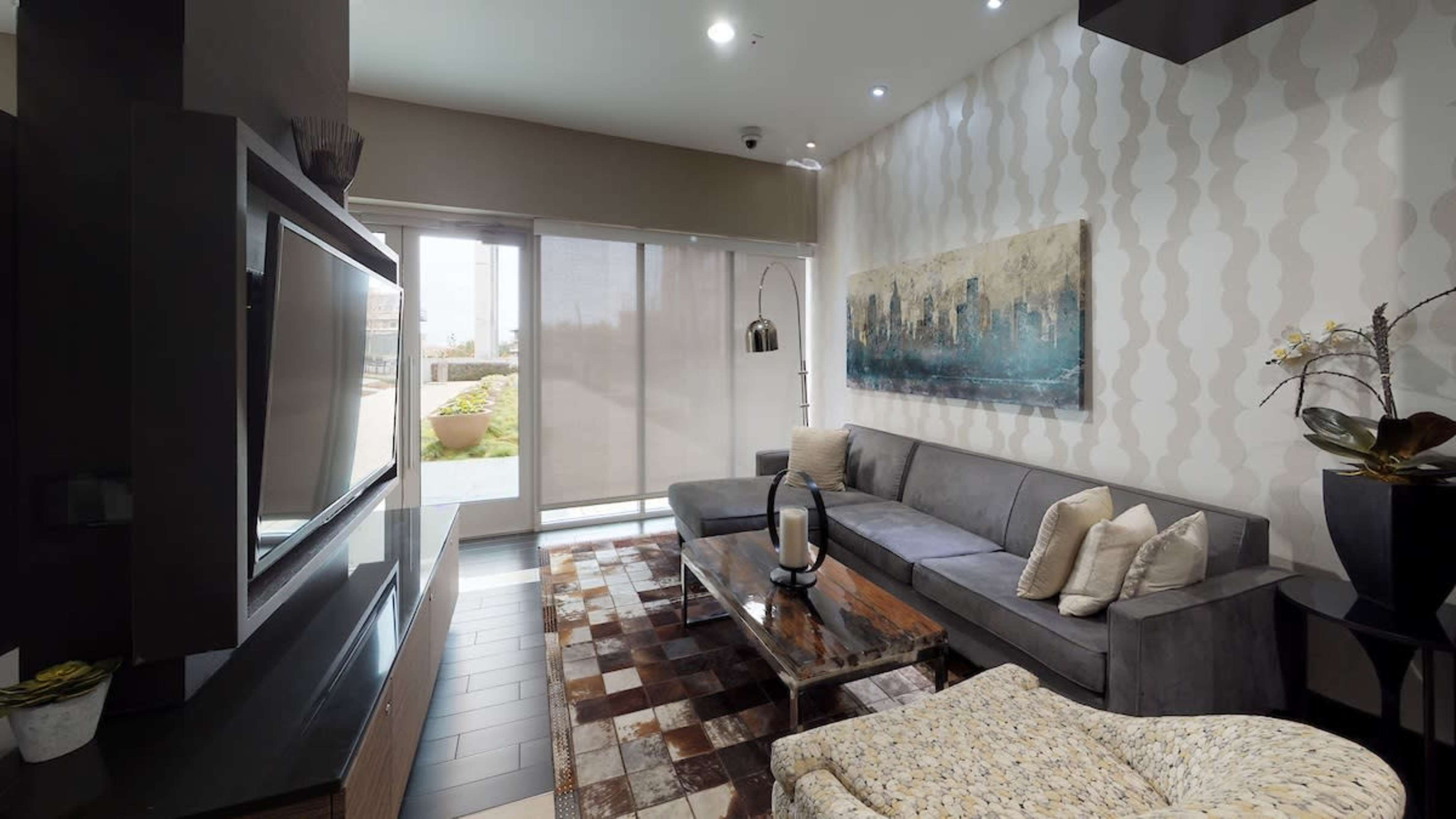 A modern living room features a gray sofa, a wooden coffee table, and large windows with blinds, overlooking a landscaped area.
