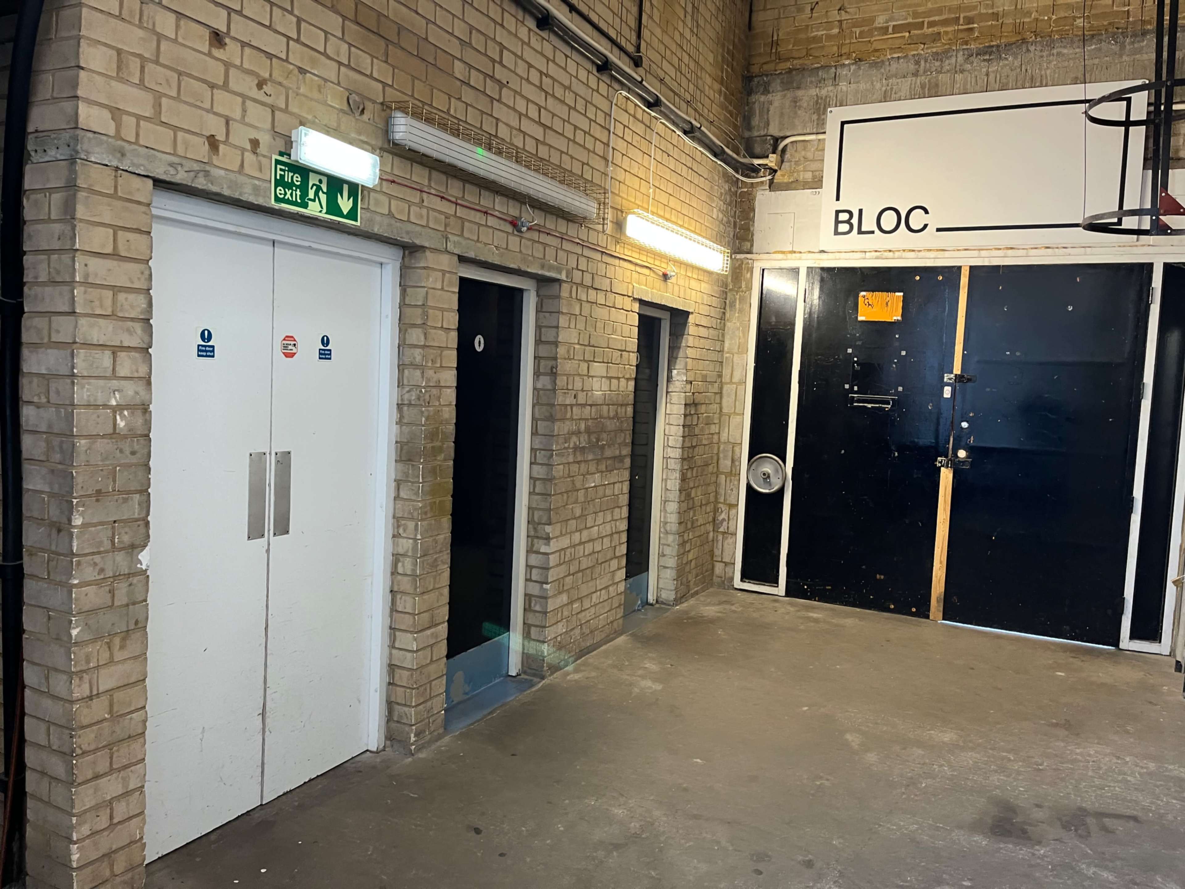 The image shows a corridor with two doors on the left side and a large black door on the right, all set against a brick wall.