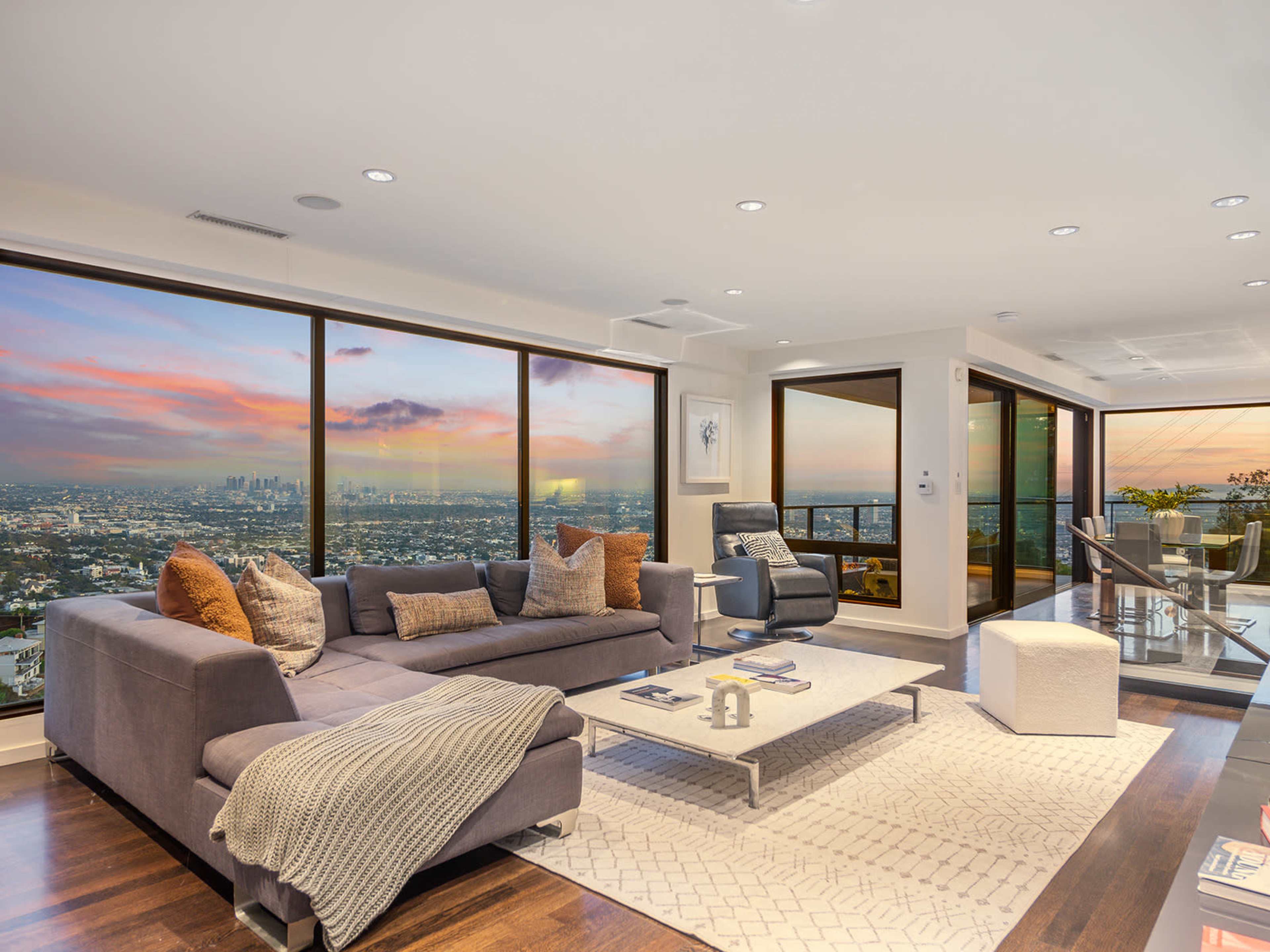 A modern living room features a gray sectional sofa, a coffee table, and large windows with a view of a sunset over the city skyline.