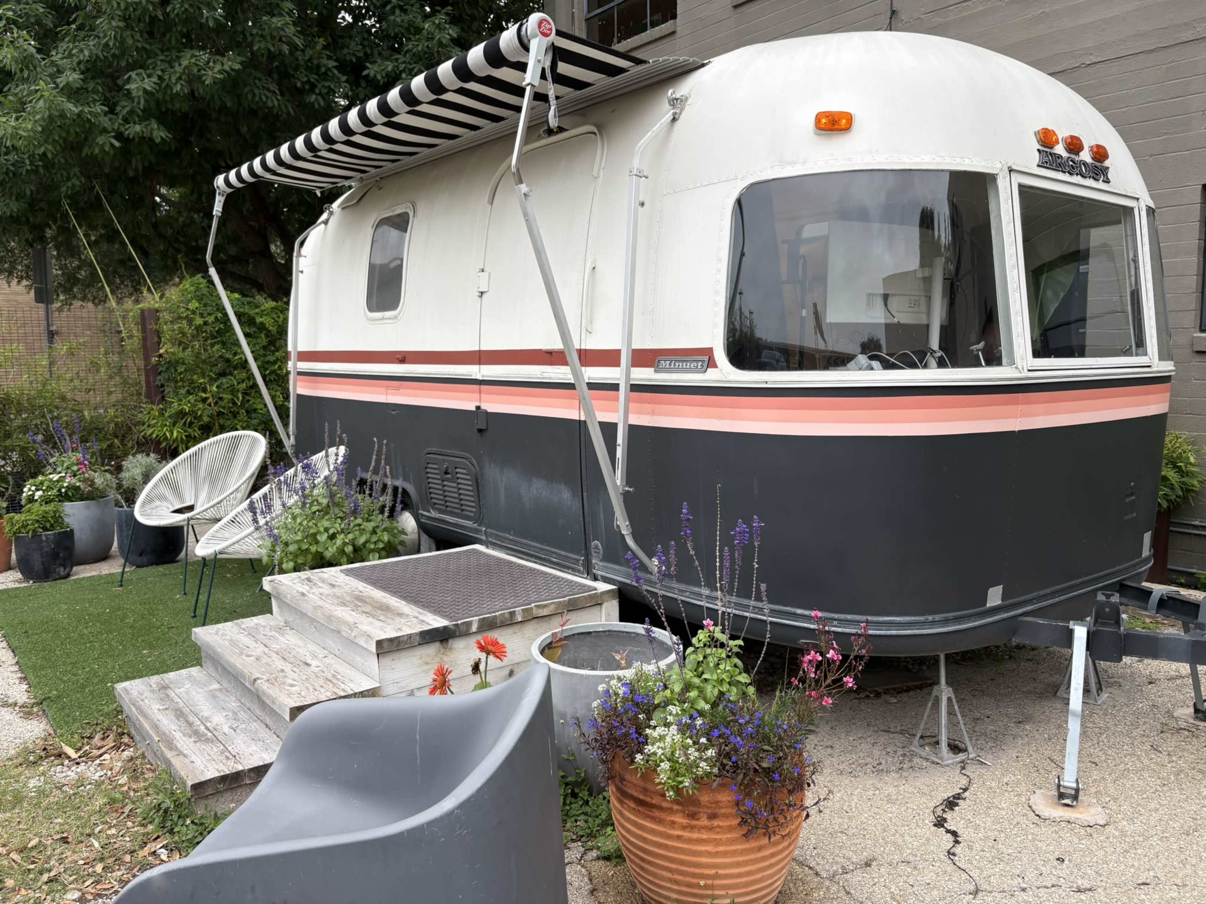 A vintage travel trailer with a striped awning is parked beside a small outdoor seating area filled with various potted plants.