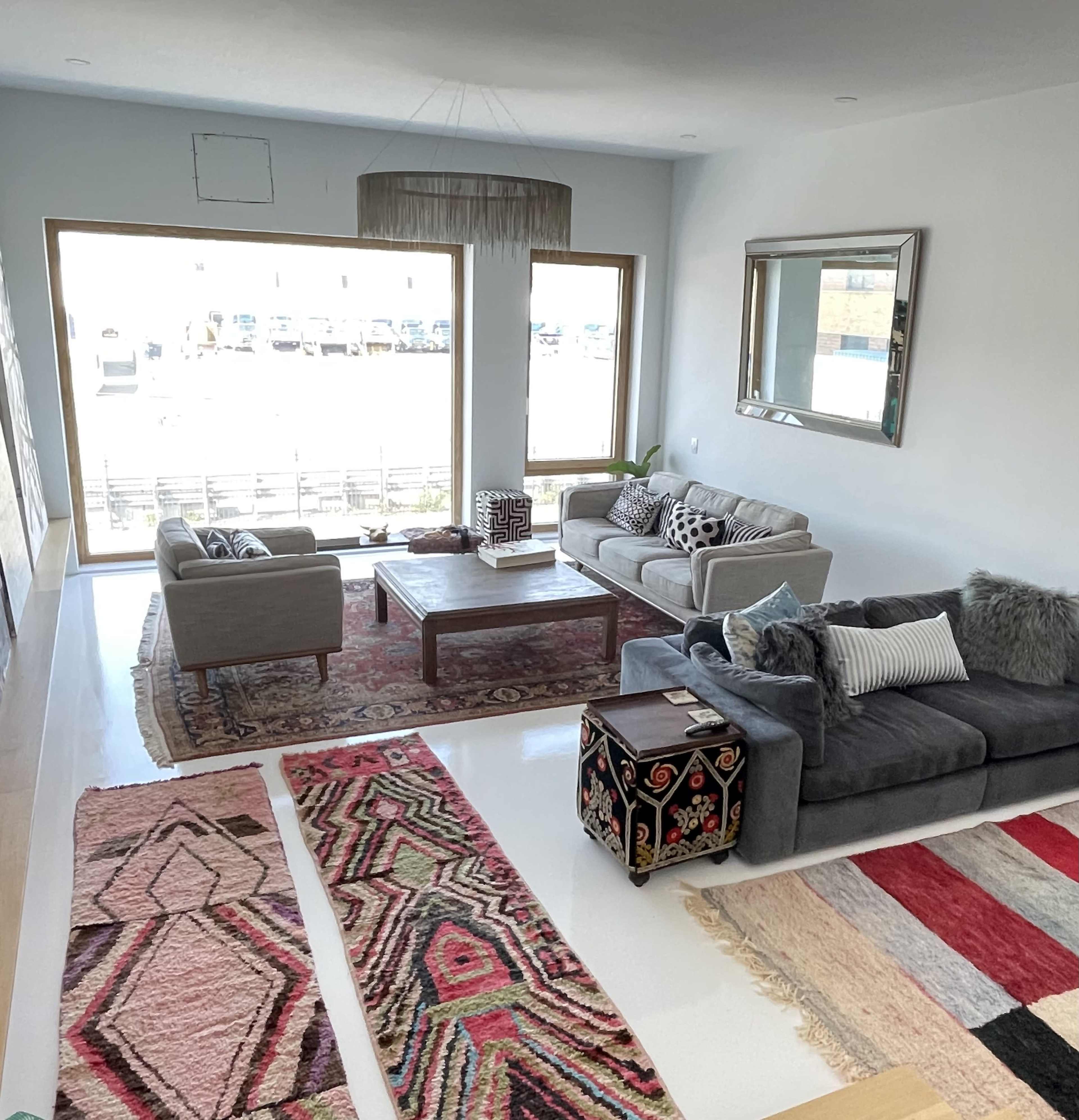 The spacious living room features two sofas, a coffee table, and a variety of patterned rugs, with large windows allowing natural light to fill the space.