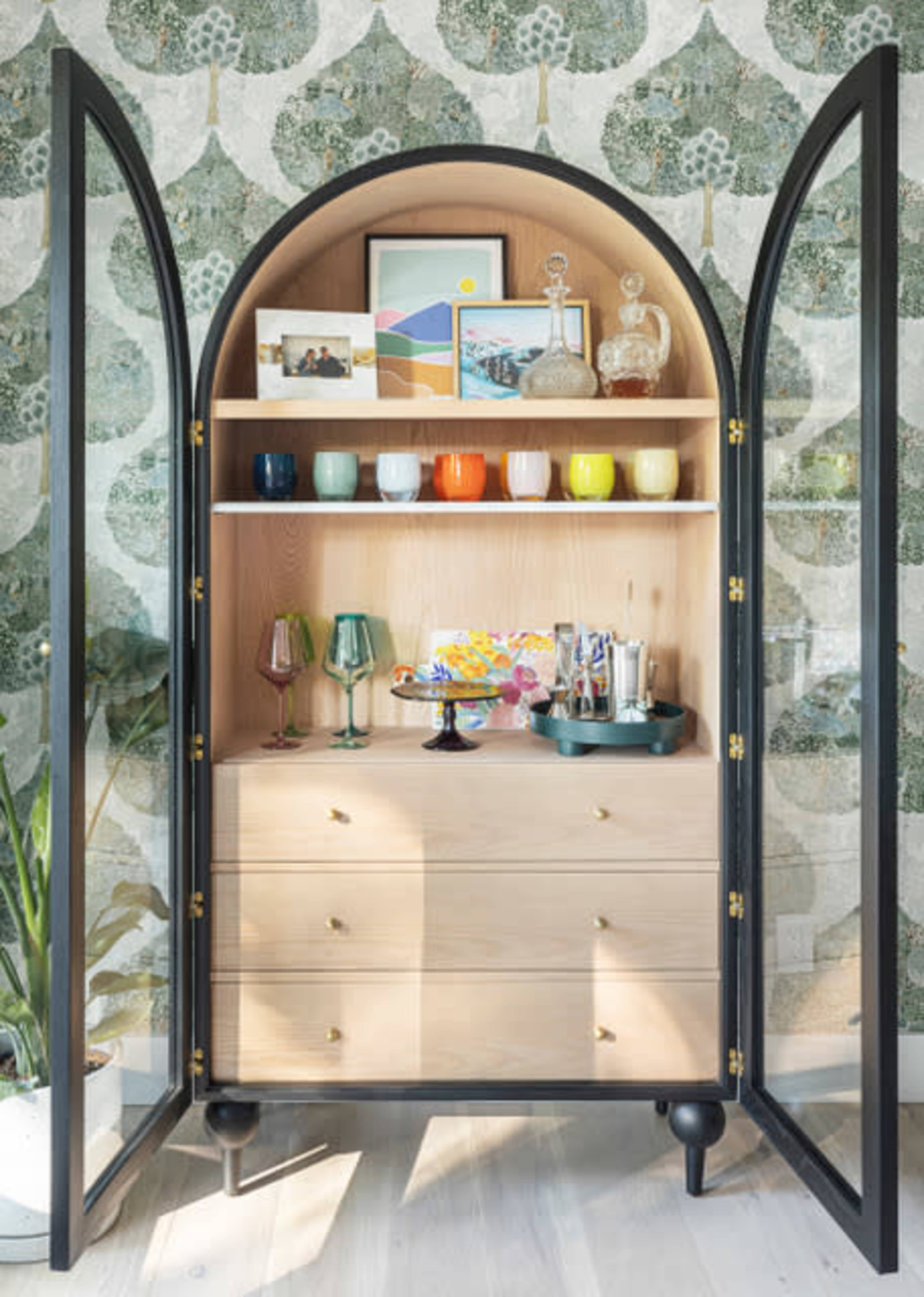 A neatly organized cabinet displays colorful glassware and decorative items against a patterned wallpaper backdrop.