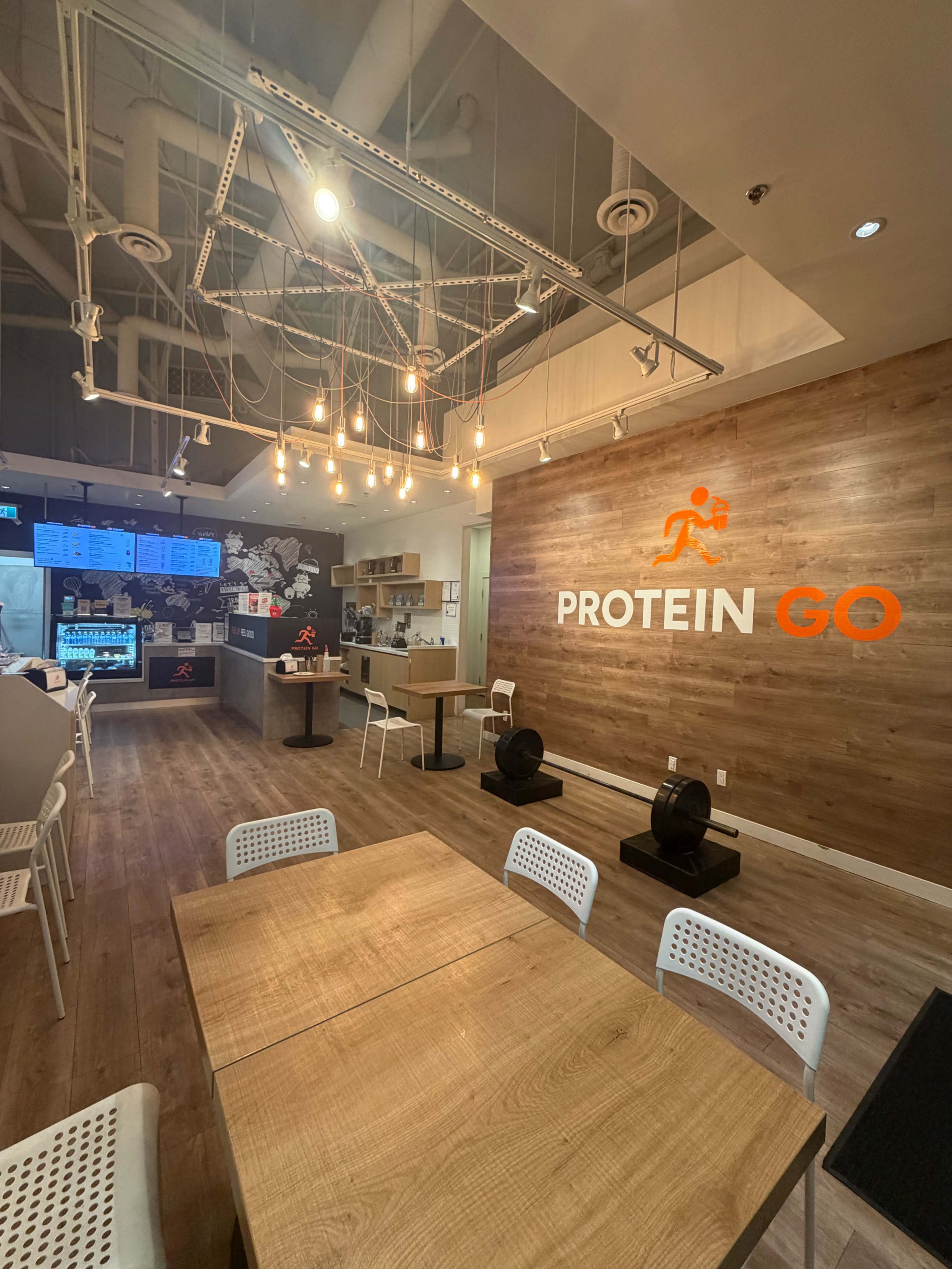 The image shows a modern interior of a protein-focused café featuring wooden accents, a service counter with menus displayed, and white tables with chairs.
