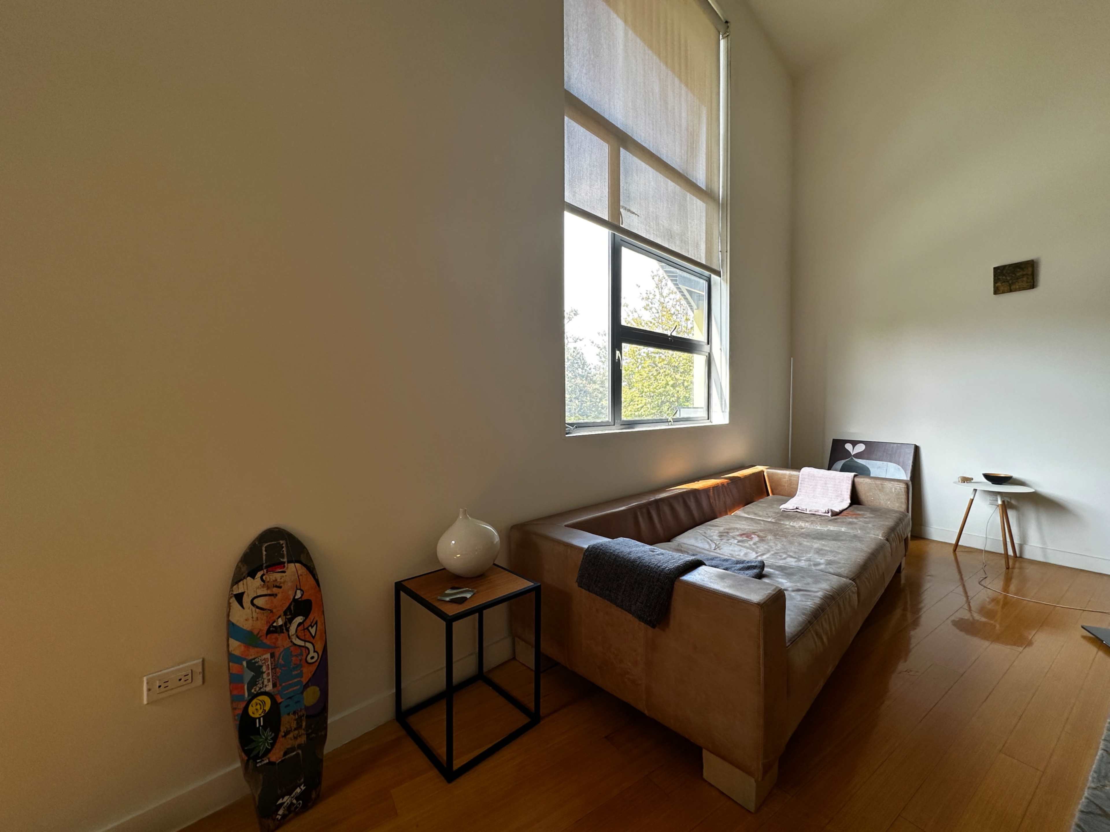 A minimalist room features a light-colored couch, a small side table, a skateboard against the wall, and a large window allowing natural light to enter.