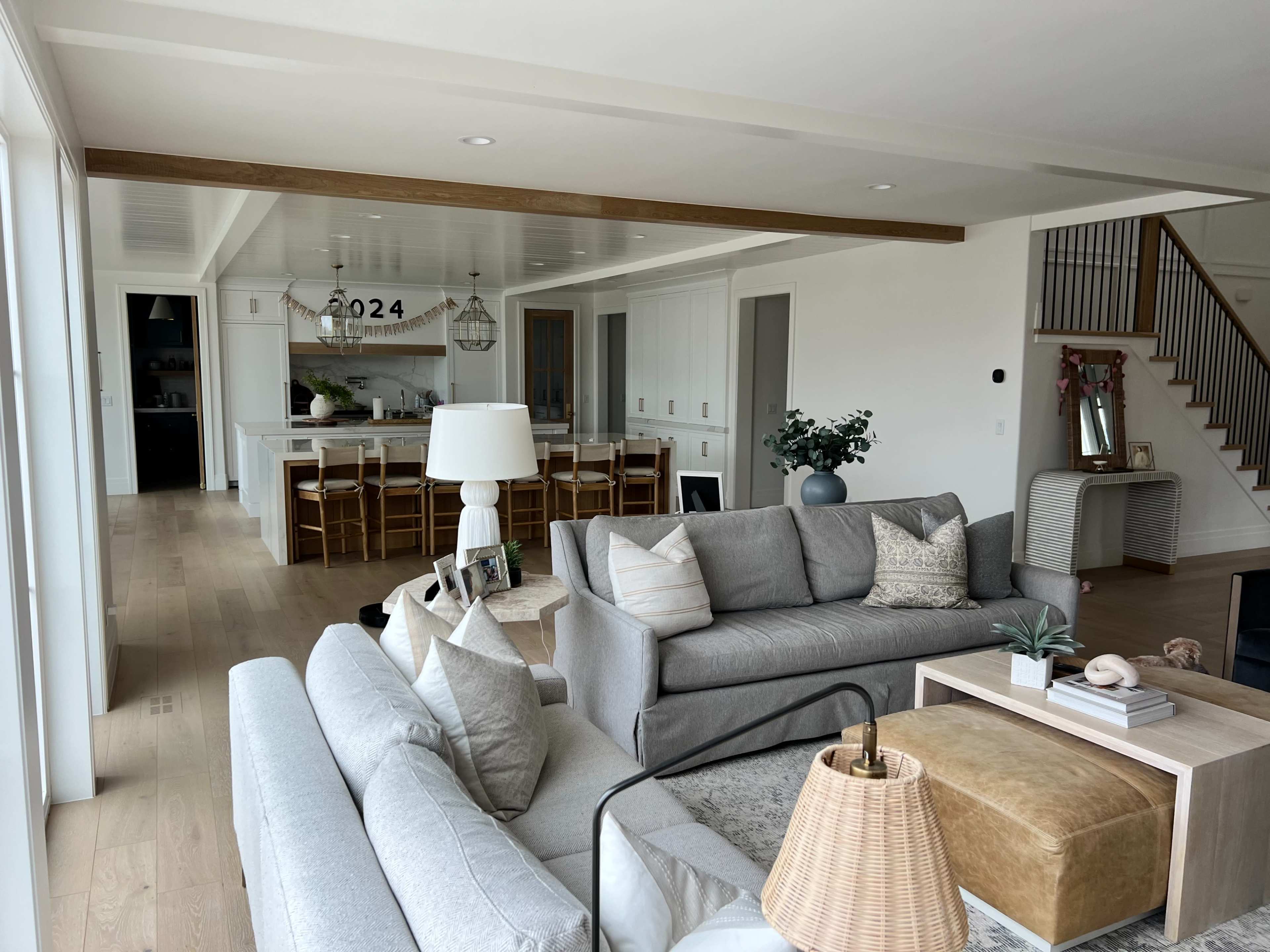 A modern living room with a light-colored sofa, coffee table, and dining area visible in the background, featuring a spacious kitchen and staircase.