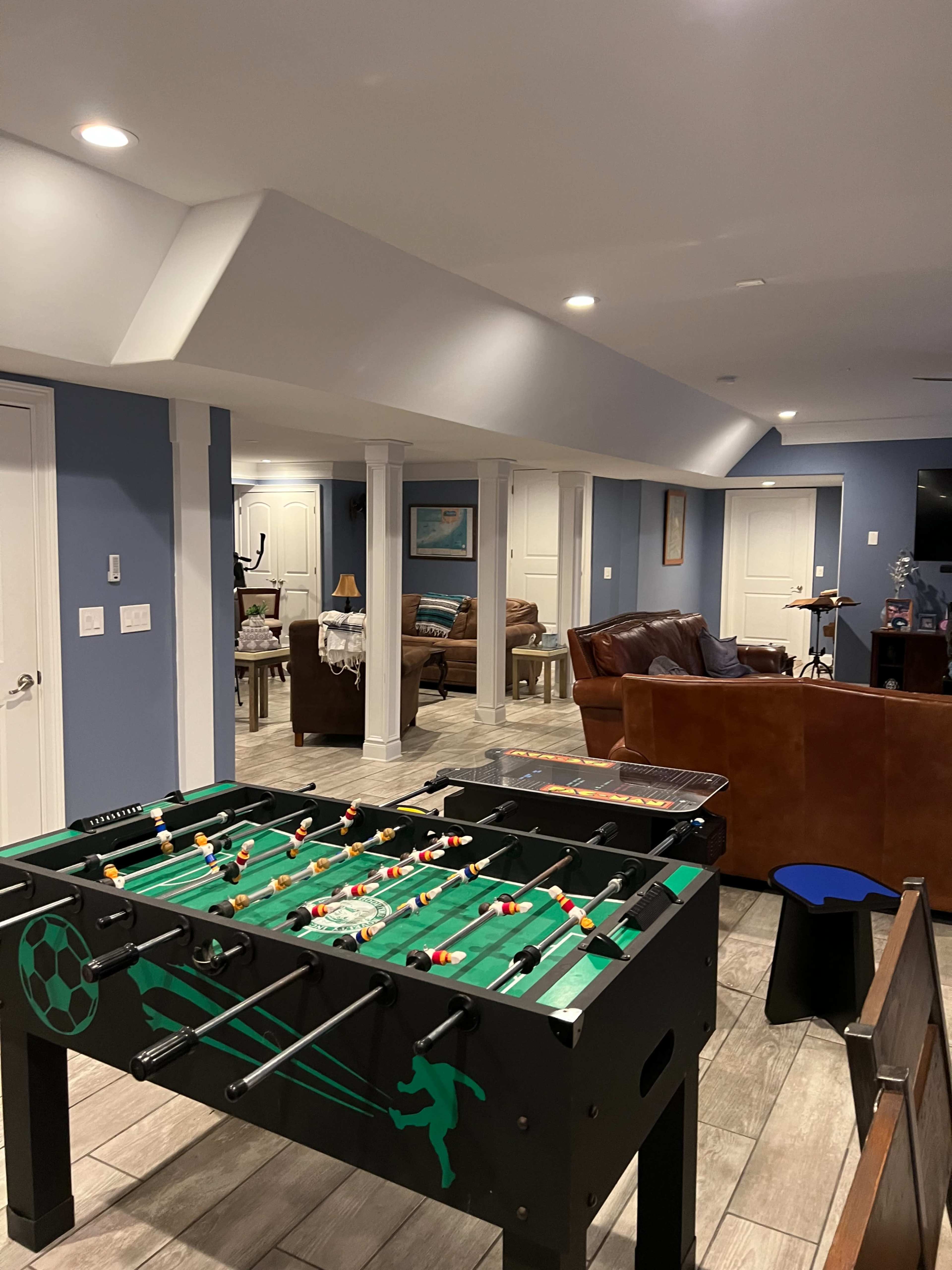 A basement game room featuring a foosball table in the foreground, with a couch and additional seating in the background.