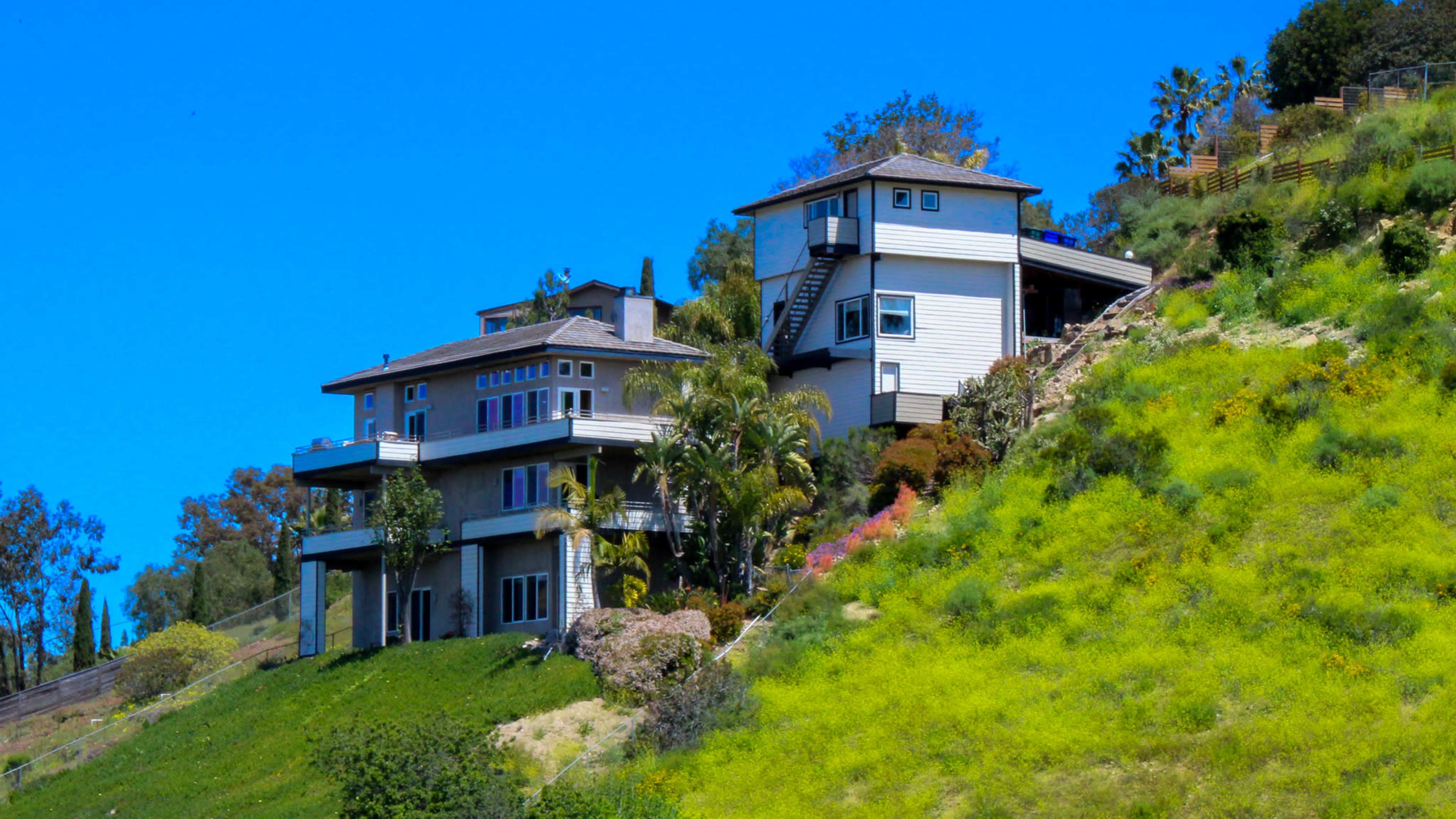 Two multi-level houses sit on a hillside covered with green grass and shrubs under a clear blue sky.