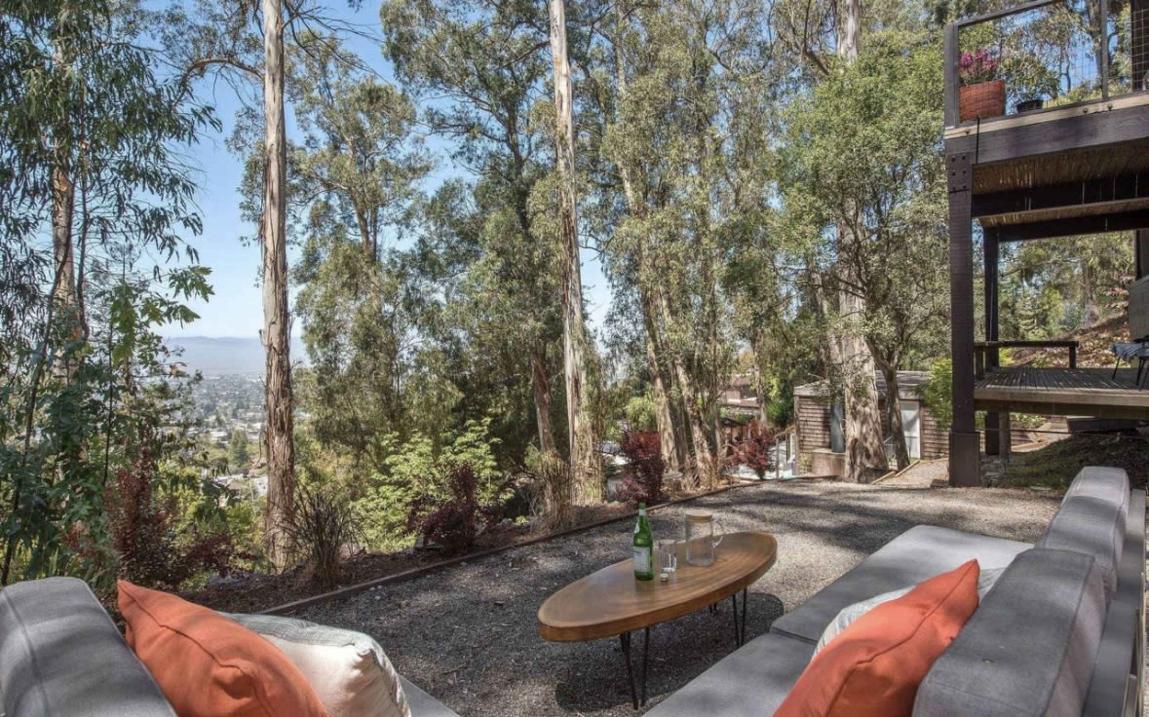 A outdoor seating area with a wooden table and two glasses sits amidst tall eucalyptus trees, overlooking a valley.