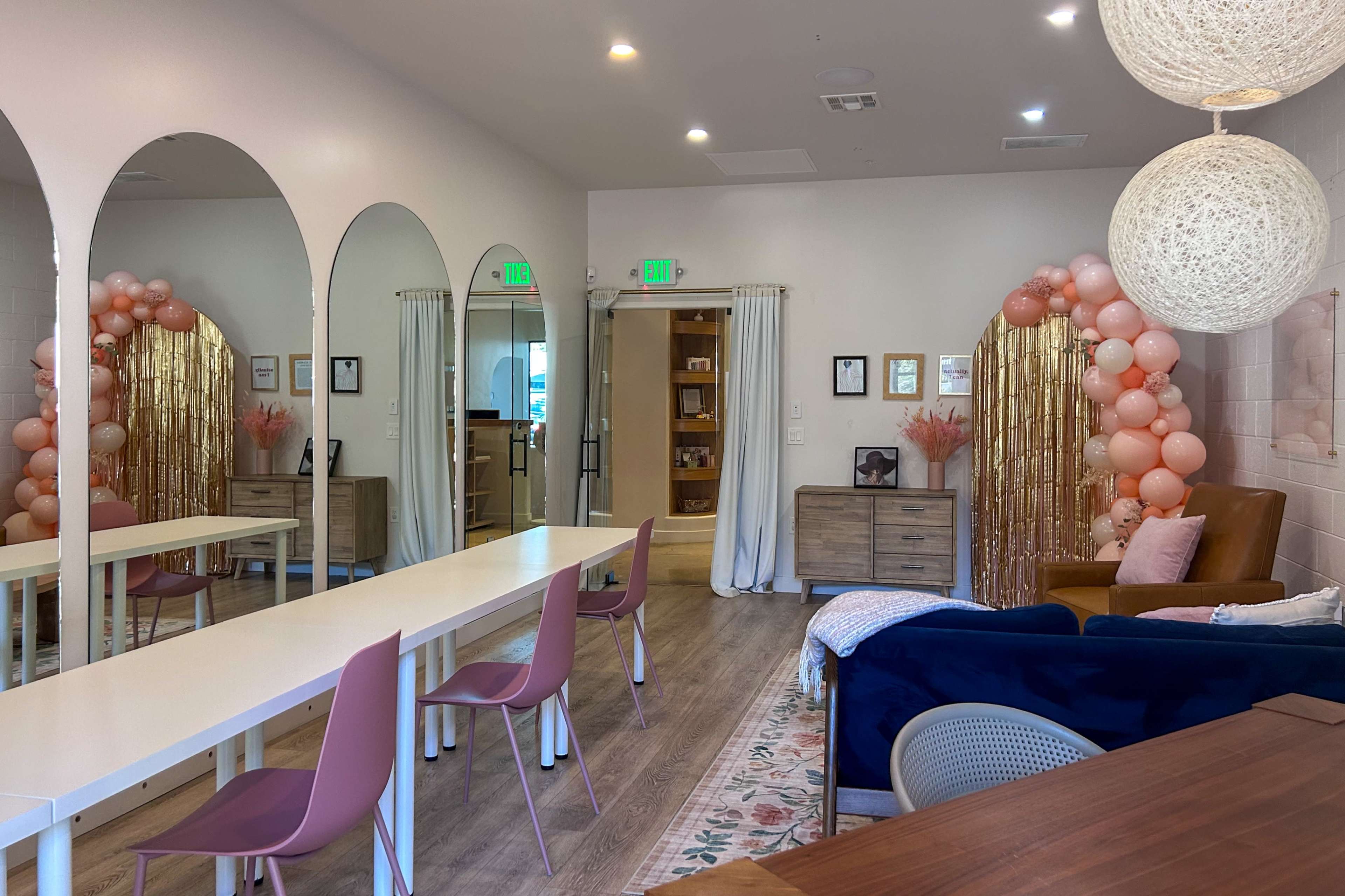 The image shows a brightly lit room with a long white table, pastel chairs, and a decorative wall featuring arches and a balloon arrangement.