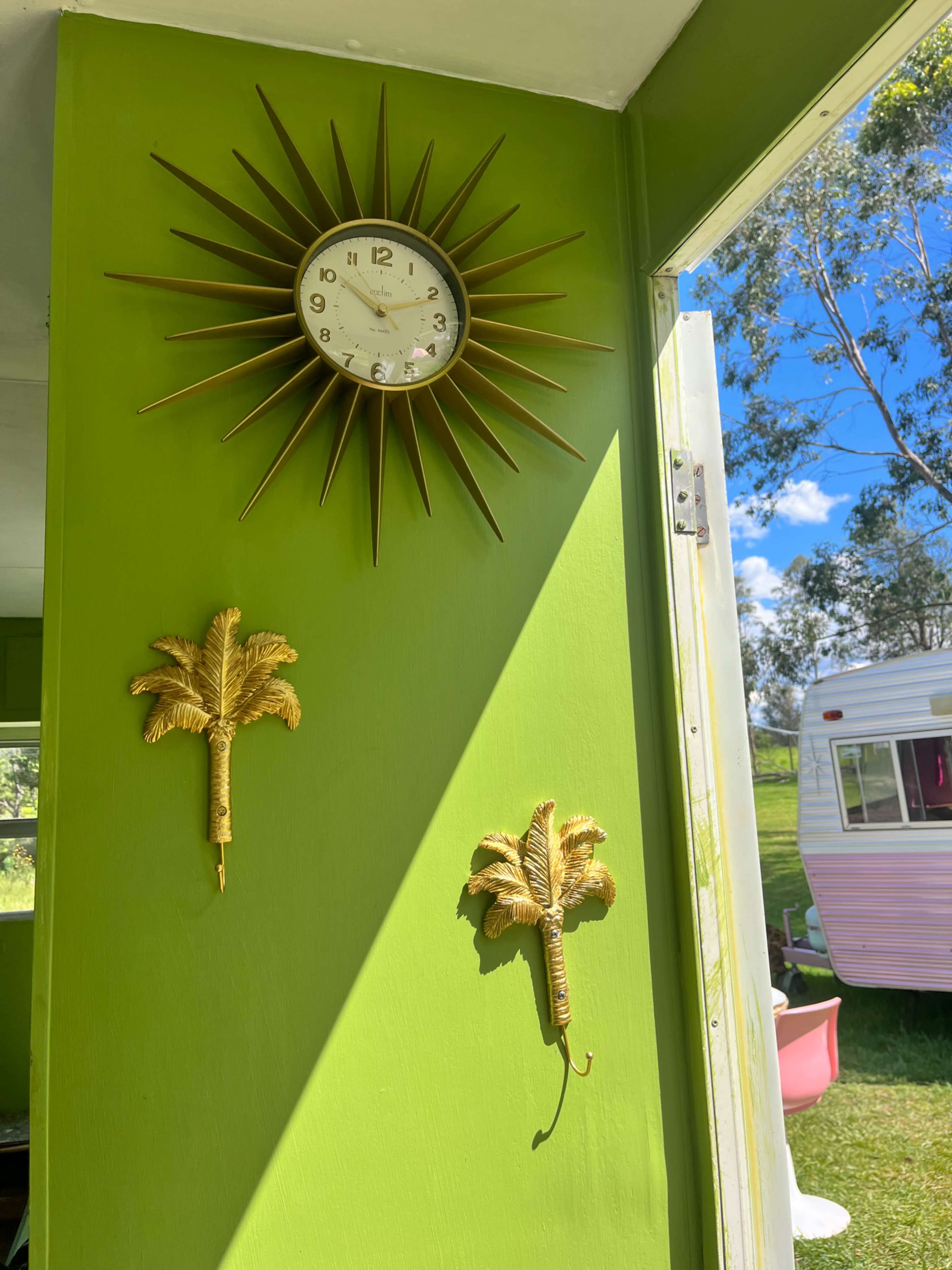 The image shows a vibrant green wall featuring a gold sunburst clock and two palm tree wall hooks, with a view of a blue sky and trees visible through an open door.