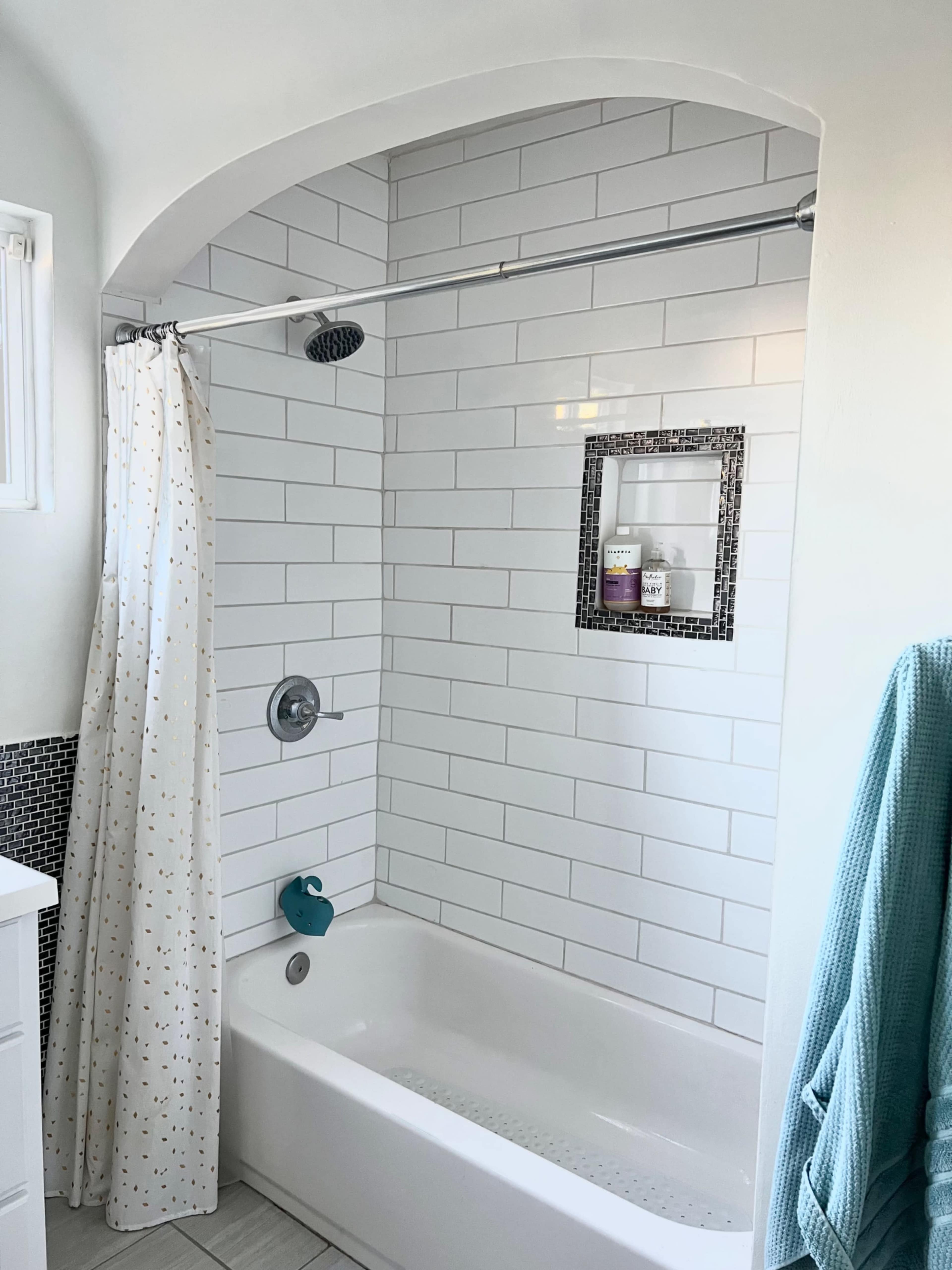 This image shows a bathroom with a tiled shower area, a bathtub, a curved shower rod, and a shelf displaying toiletries.