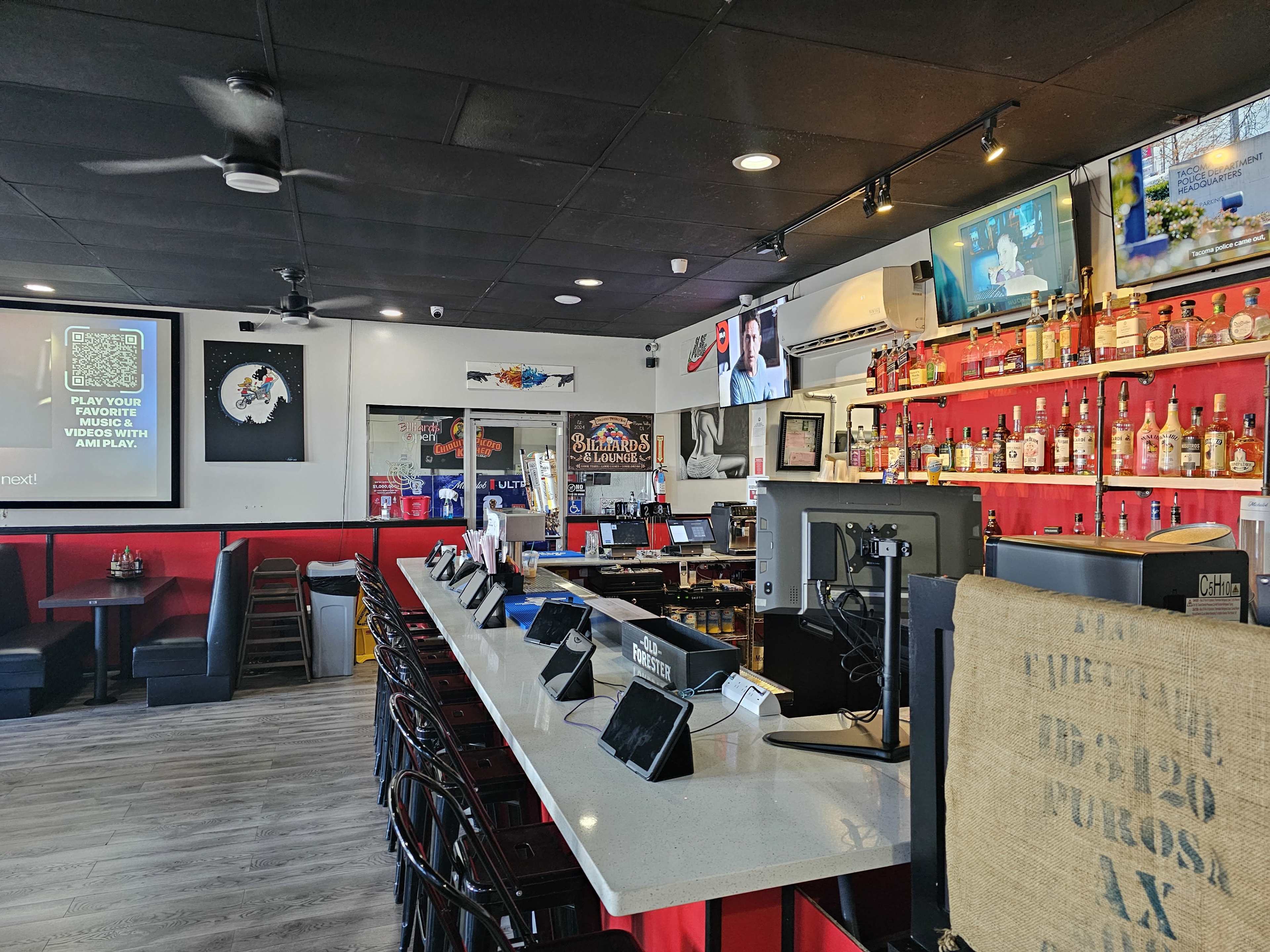 The image shows an interior view of a bar with a long, sleek counter, multiple liquor shelves, and a screen displaying a QR code on the wall.